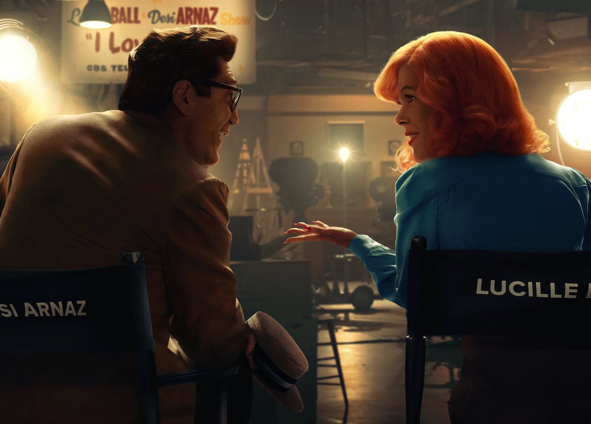 Javier Bardem and Nicole Kidman pose as Desi Arnaz and Lucille Ball in the 'Being the Ricardos' poster. Their backs and profiles are facing the camera as they talk while seated on the fictional set of 'I Love Lucy' made for the Aaron Sorkin film.
