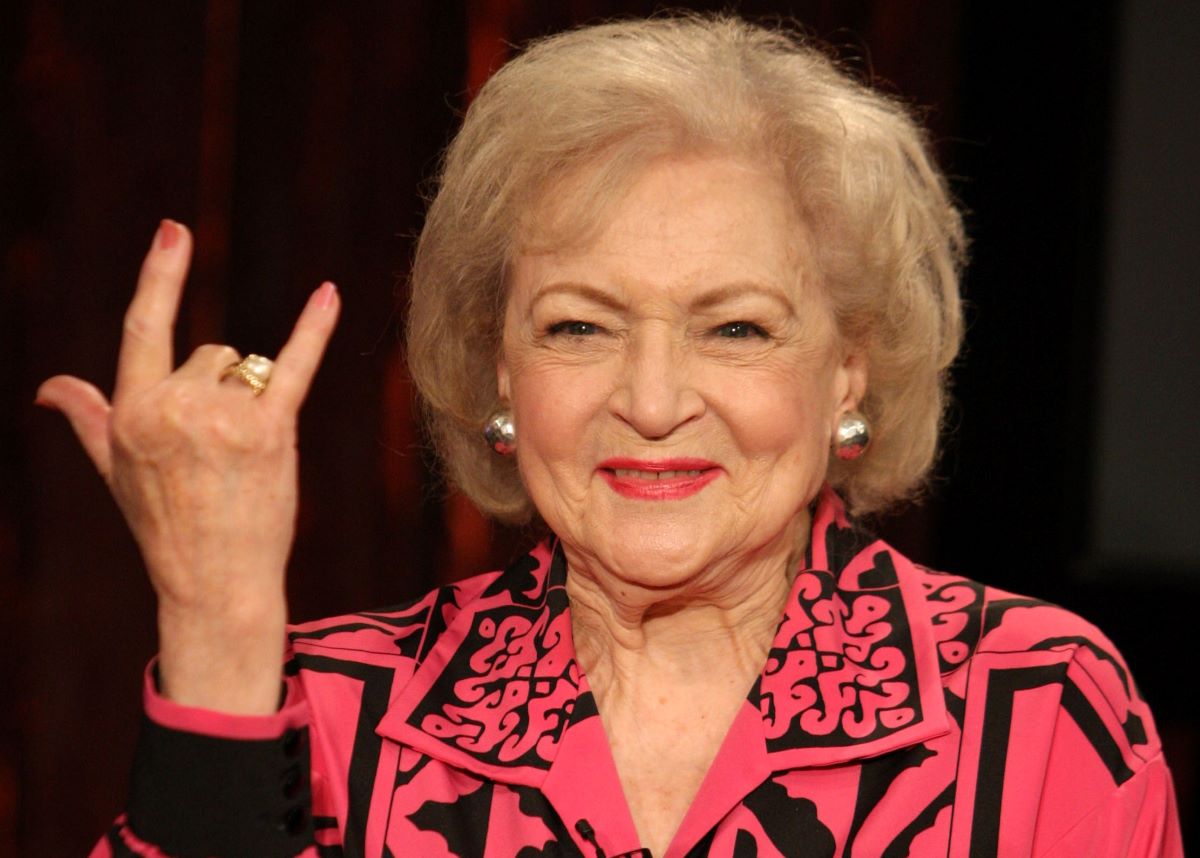 Betty White in a pink shirt, smiling and flashing a 'rock on' hand sign