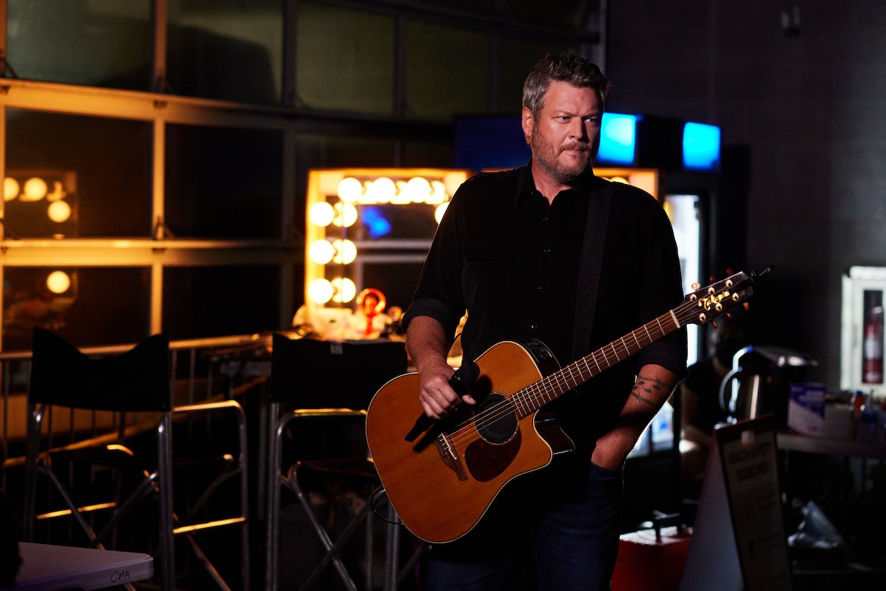 Blake Shelton stands with a guitar and holds a microphone.