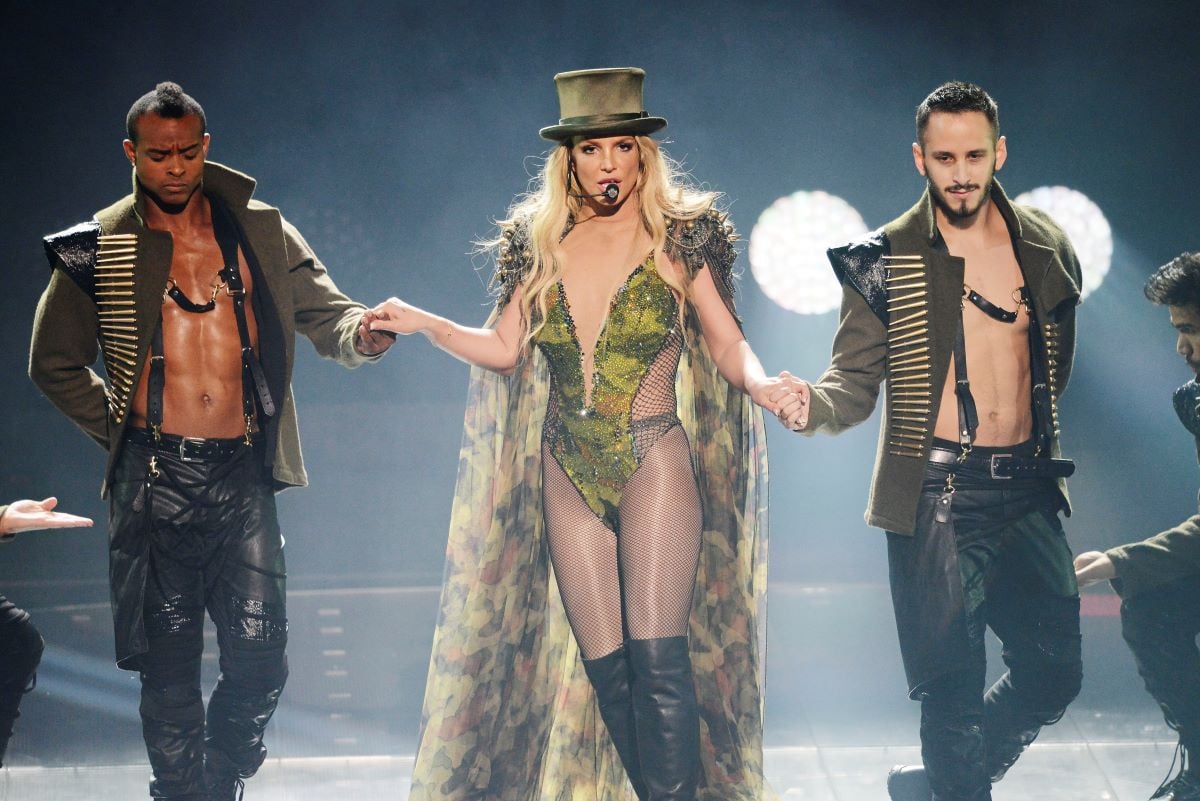 Britney Spears dressed in a body suit, cape, and top hat, with dancers on either side of her