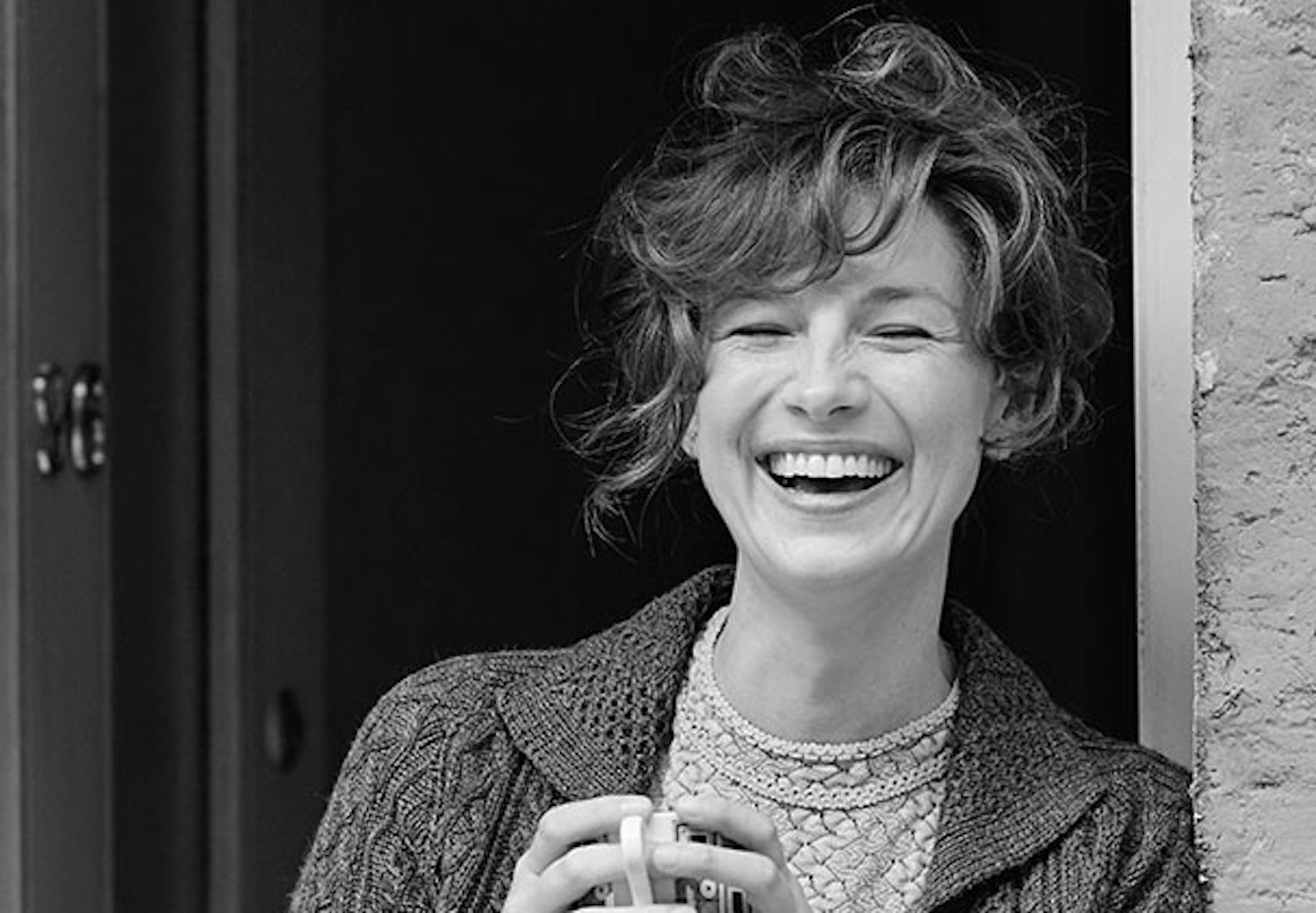 Caitriona Balfe in 'Belfast.' In this black-and-white still from the Kenneth Branagh movie, Balfe stands in a doorway laughing with a teacup in her hands. She wears two sweaters and her hair is curly and up on her head.