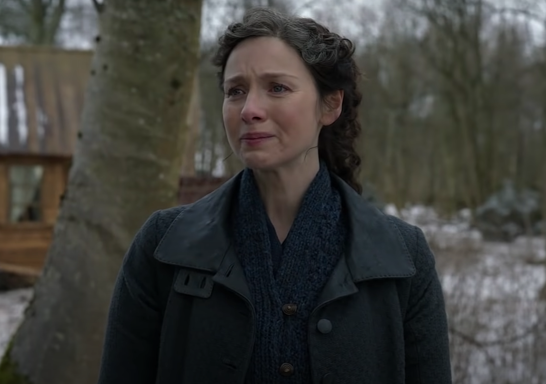 Caitriona Balfe in the 'Outlander' Season 6 trailer. She stands in a navy blue sweater and dark grey coat holding back tears. Surrounding her is a snowy forest and a cabin in the background.