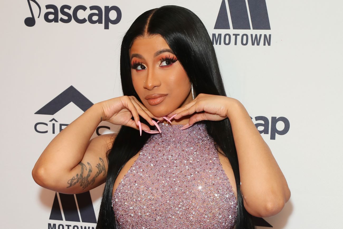 Cardi B wears a purple sequined dress. She holds her hands near her face.