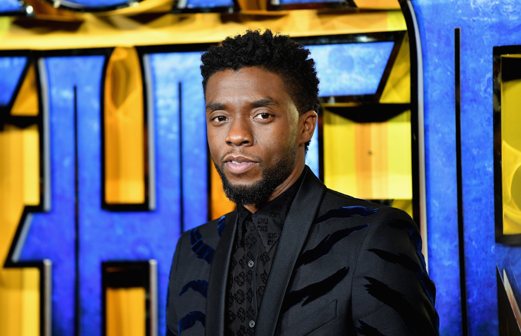 'Black Panther' star Chadwick Boseman. He's wearing a black suit and standing in front of a blue and yellow wall.