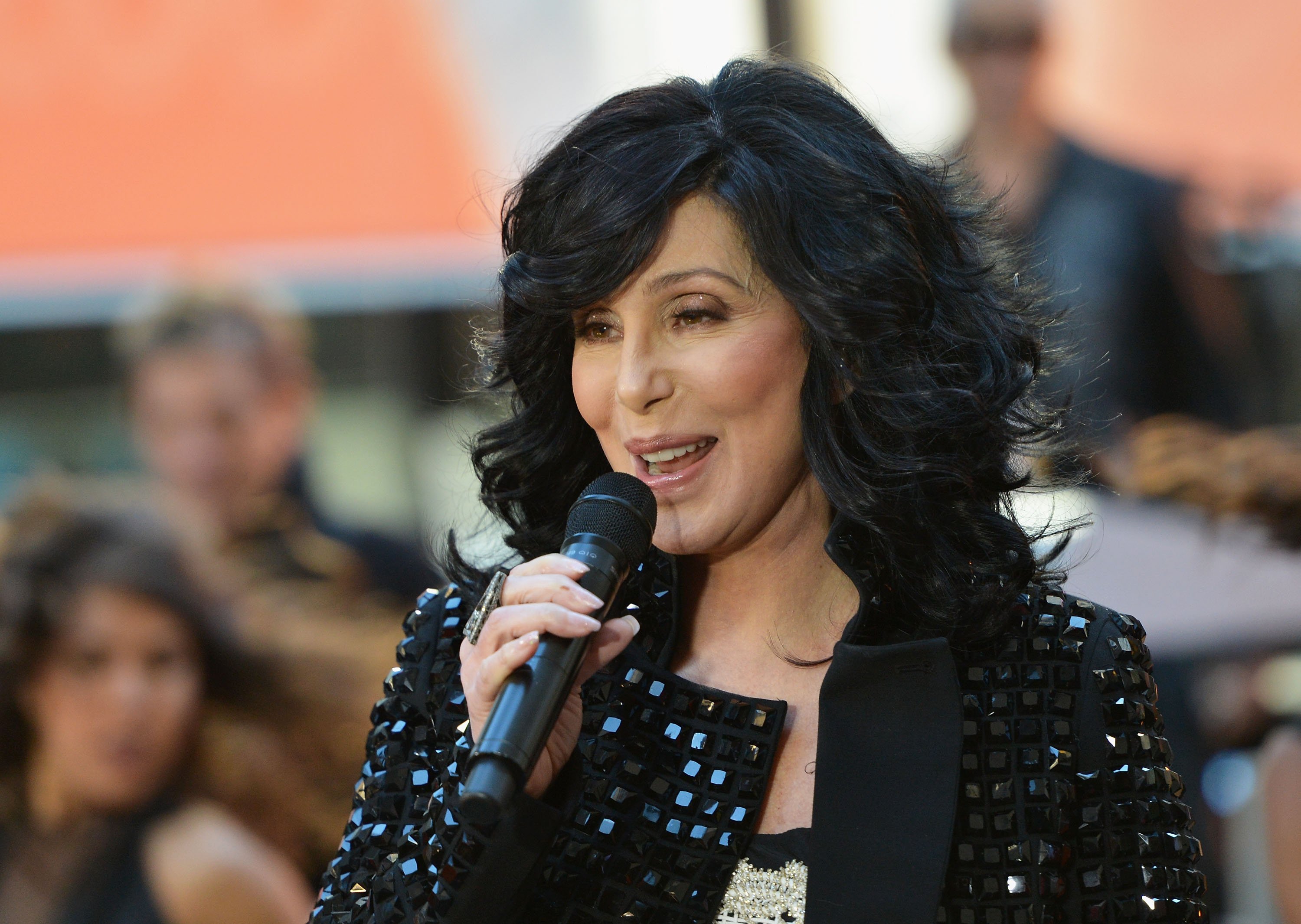 Cher wears a black jacket and speaks into a microphone.