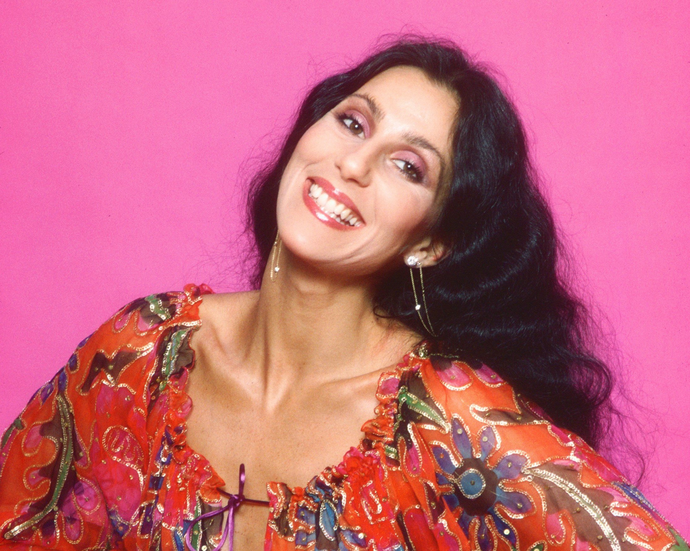 Cher wears a red floral shirt and stands in front of a pink background.