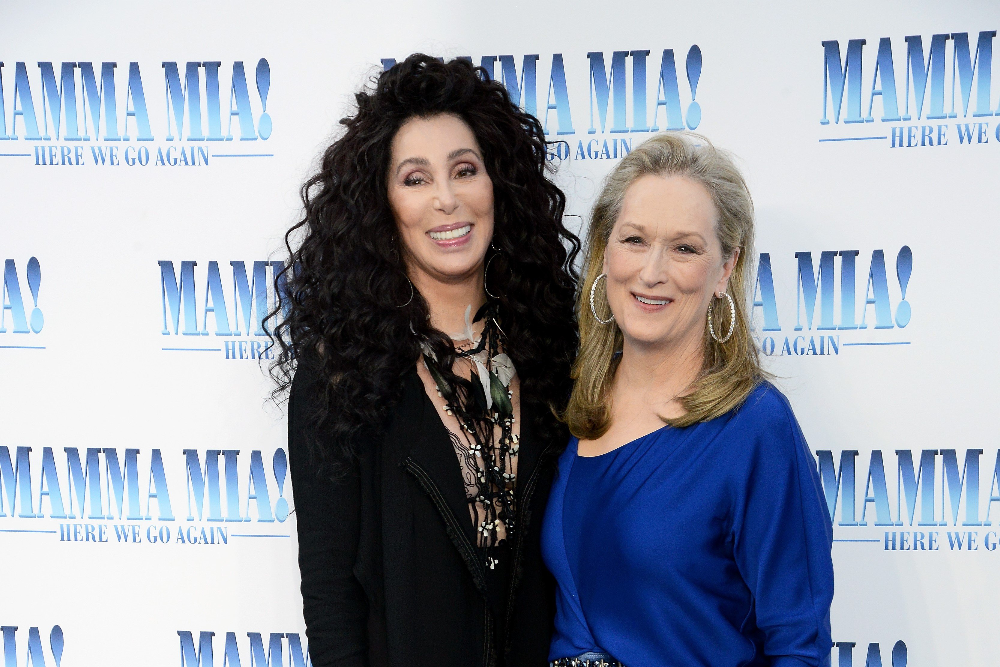 Cher wears a black dress and Meryl Streep wears a blue dress. They stand in front of a 'Mamma Mia! Here We Go Again' poster.