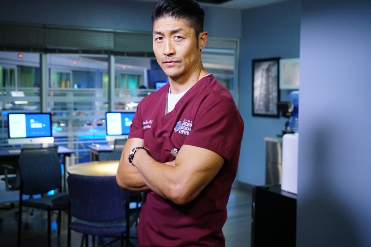 Brian Tee as Dr. Ethan Choi returns to 'Chicago Med' Season 7. Dr. Choi is wearing maroon scrubs and has his arms crossed.