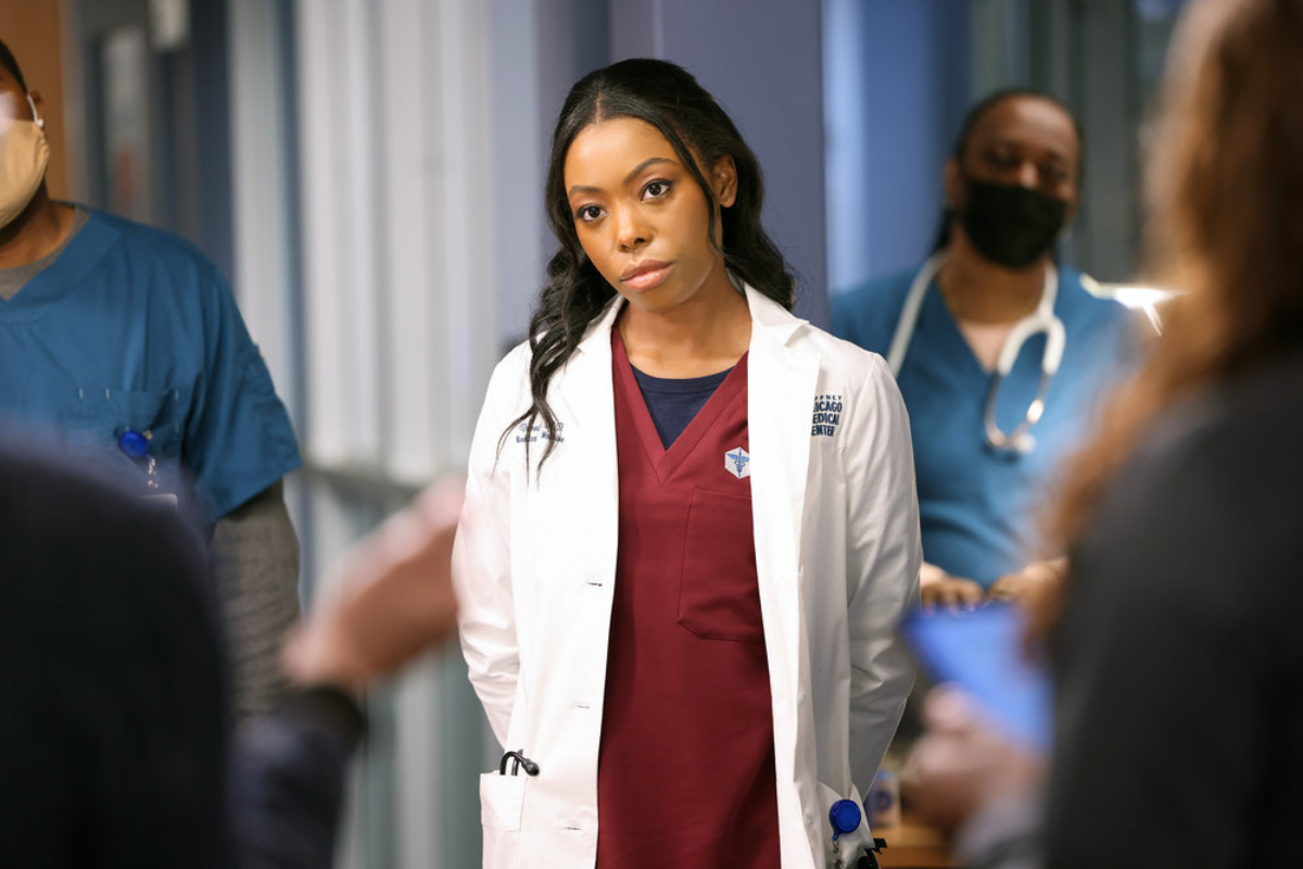 Asjha Cooper as Vanessa Taylor in Chicago Med Season 7 Episode 8. Vanessa is wearing maroon scrubs and a white coat and appears to be listening to someone. 