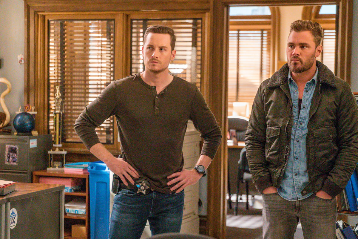 Jesse Lee Soffer as Jay Halstead and Patrick John Flueger as Adam Ruzek in Chicago P.D. Halstead and Ruzek stand next to each other. Halstead wears a brown long-sleeved shirt and Ruzek wears a denim button-up and jacket.