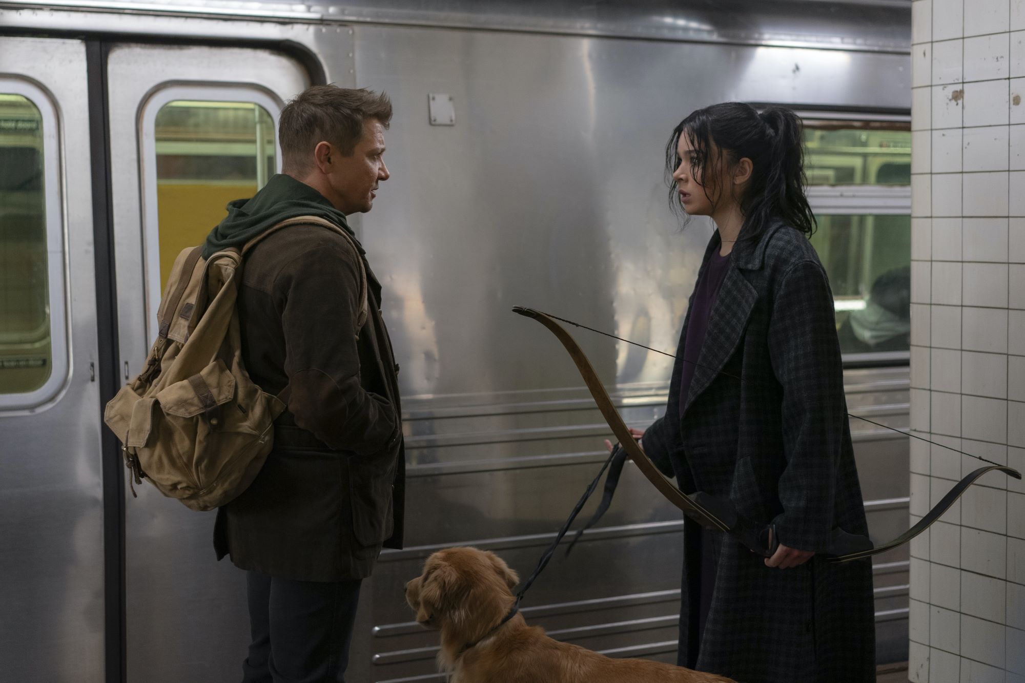 'Hawkeye' stars Jeremy Renner and Hailee Steinfeld, in character as Clint Barton and Kate Bishop, stand in a subway station as a train passes by. Clint wears a brown jacket over a green hoodie and has a tan backpack on his back. Kate wears a long dark plaid jacket over a purple shirt and is holding onto a bow in one hand and the leash that is attached to her dog in the other.