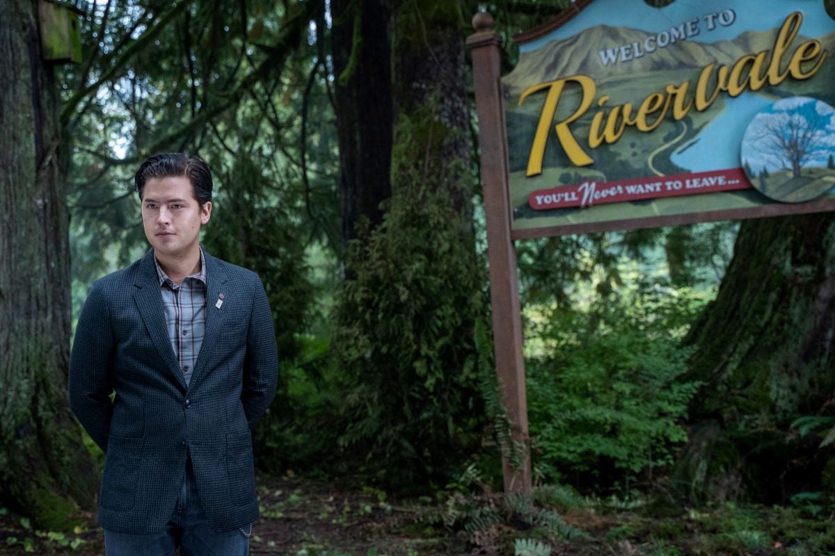 Cole Sprouse as Jughead Jones standing in front of a Rivervale town sign in Riverdale