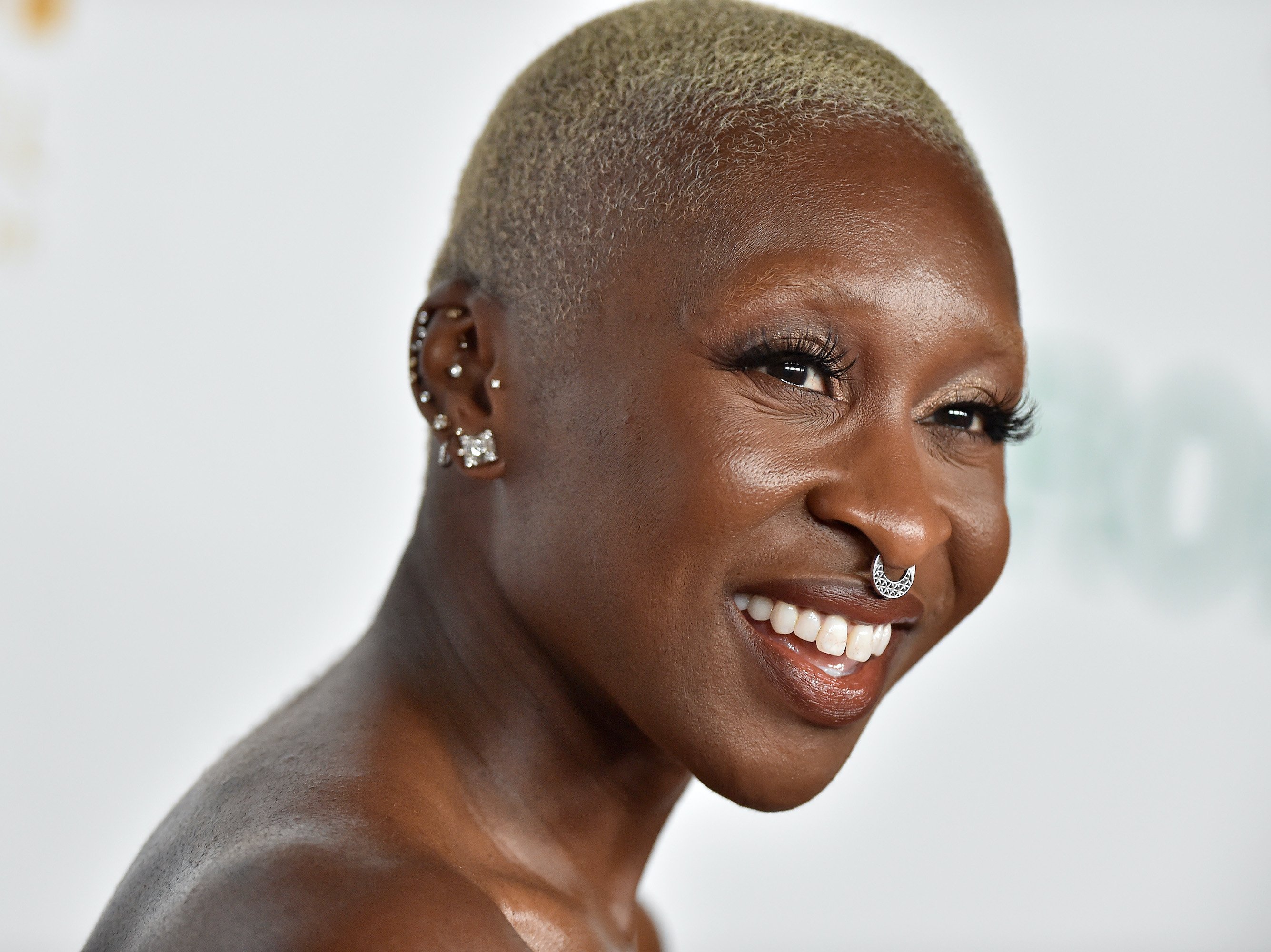 Cynthia Erivo attends the Los Angeles Philharmonic Homecoming Concert and Gala at Walt Disney Concert Hall on Oct. 09, 2021 in Los Angeles. She wears a strapless pink gown and smiles in front of a white backdrop. She has a silver septum piercing in and her ears are adorned with silver earrings all the way up.