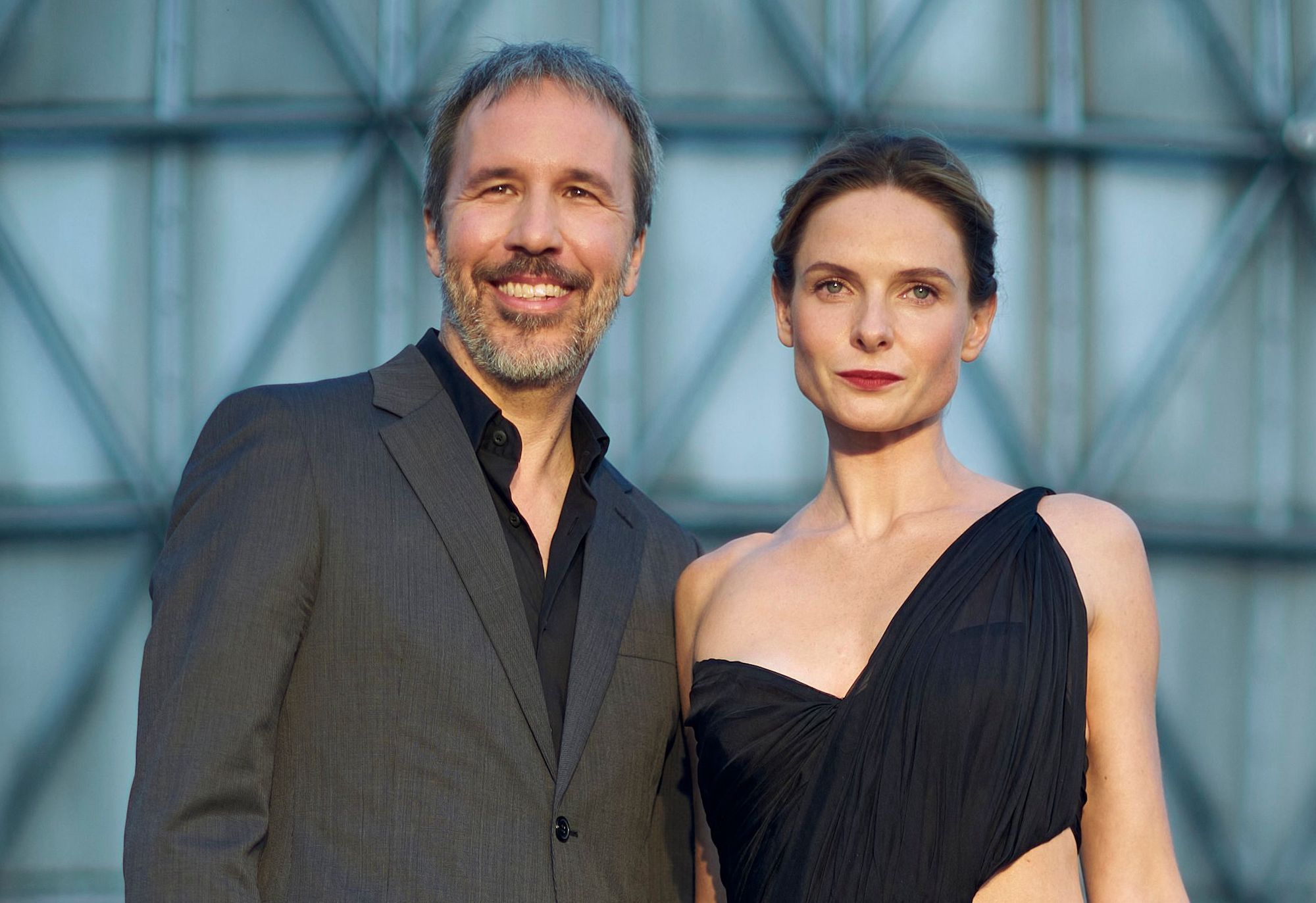 'Dune' director Denis Villeneuve and Rebecca Ferguson at the 'Dune' premiere at the Toronto International Film Festival in Toronto, Ontario, Sept. 11, 2021. They stand outside in front of a geometric backdrop. Villeneuve (L) wears a black shirt and grey suit. Ferguson (R) wears a black dress with one twisted strap on her left shoulder.