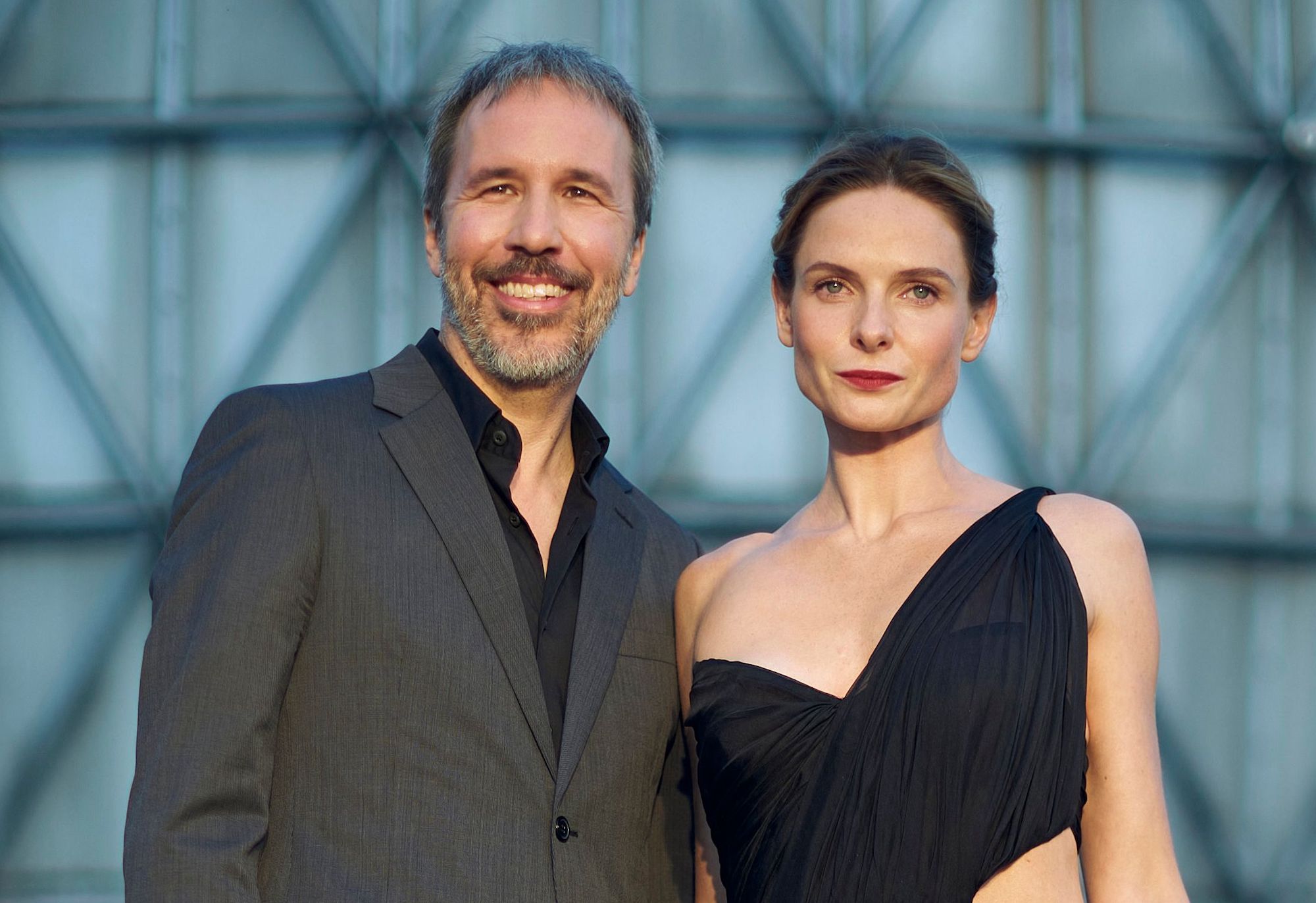 'Dune' director Denis Villeneuve and Rebecca Ferguson at the 'Dune' premiere at the Toronto International Film Festival in Toronto, Ontario, Sept. 11, 2021. They stand outside in front of a geometric backdrop. Villeneuve (L) wears a black shirt and grey suit. Ferguson (R) wears a black dress with one twisted strap on her left shoulder.