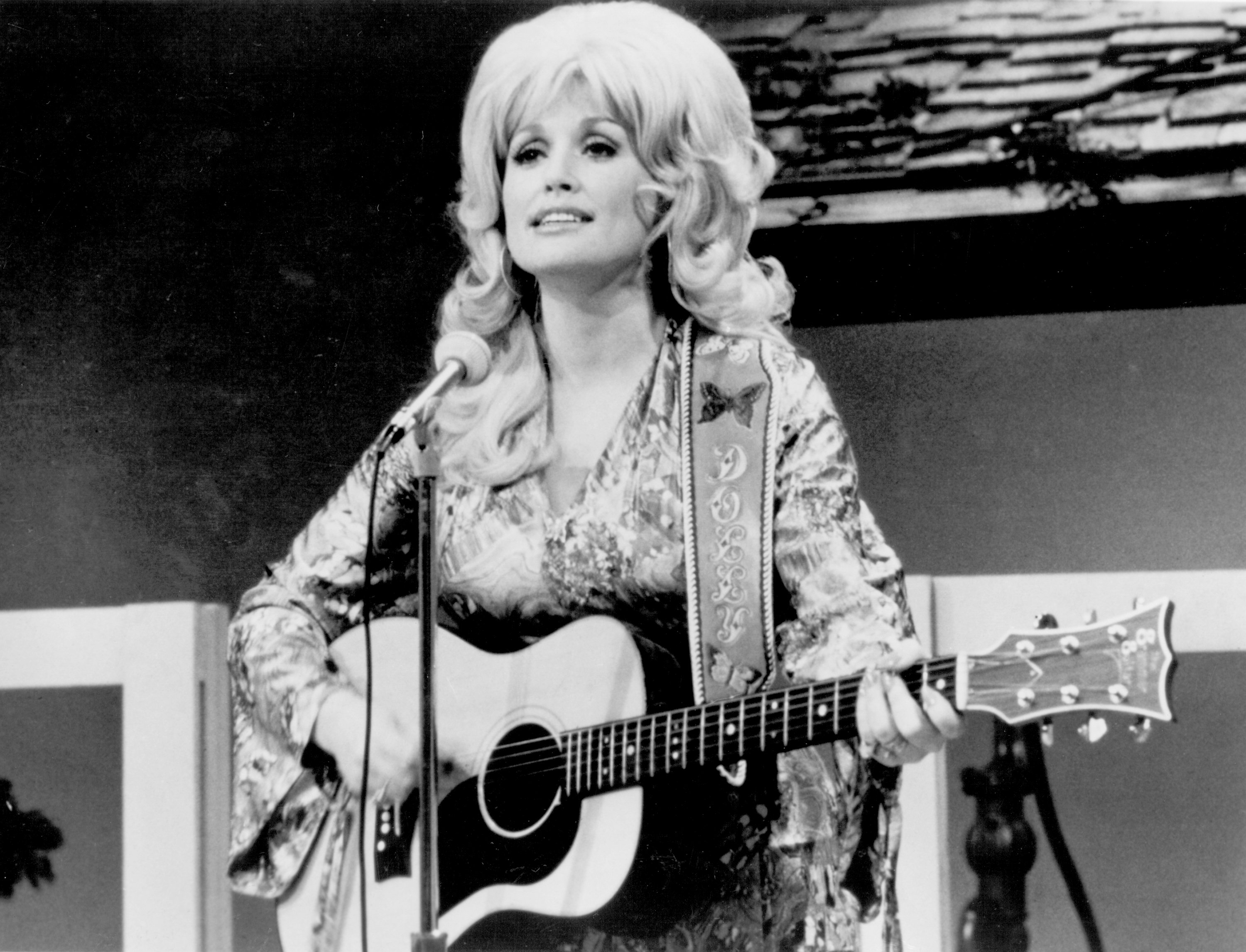 A black and white photo of Dolly Parton in a patterned shirt. She holds a guitar.