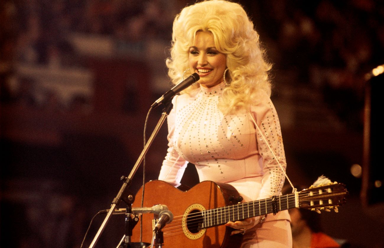 Dolly Parton wears a white rhinestoned shirt and holds a guitar. She stands in front of a microphone.