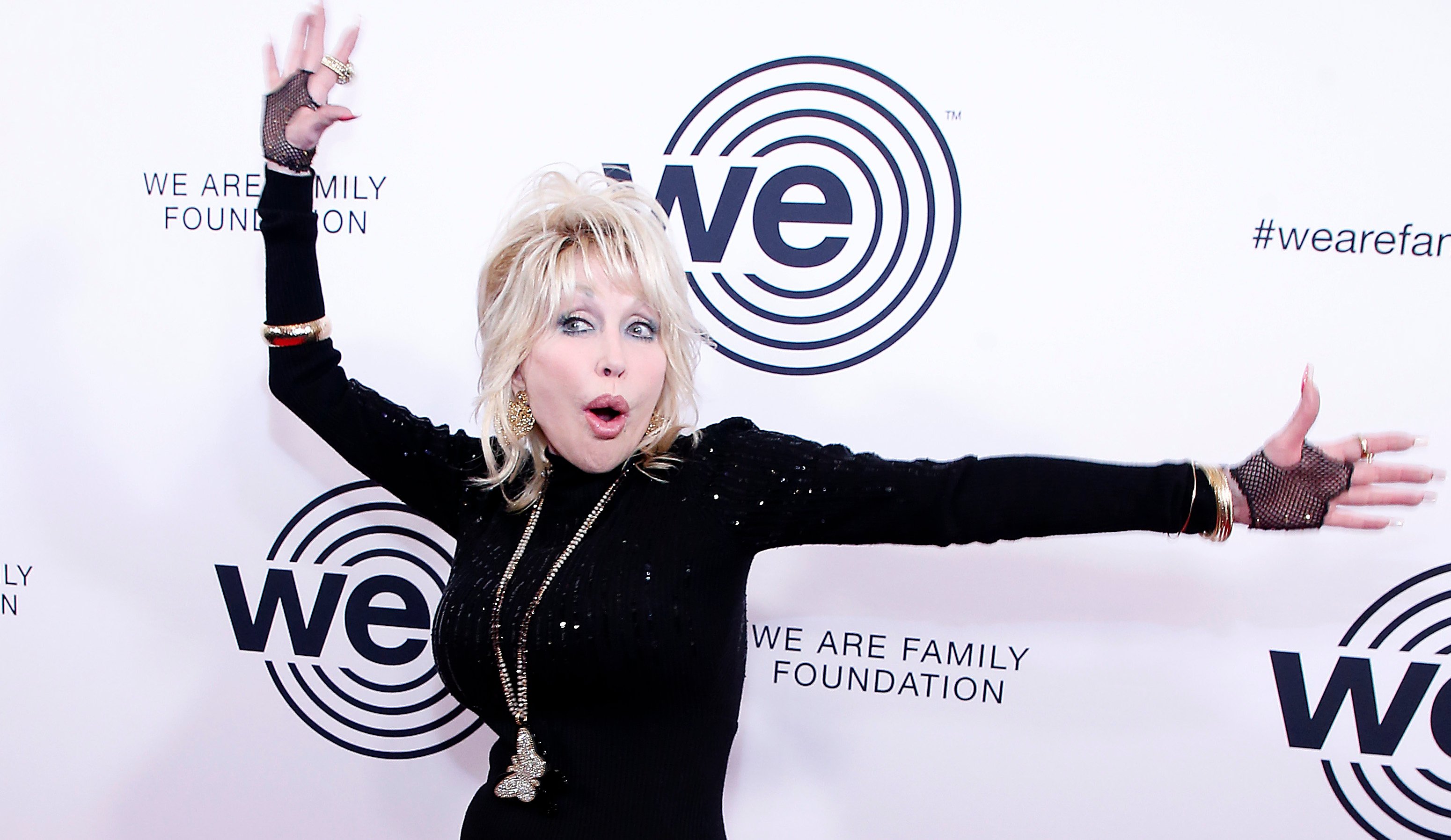 Dolly Parton poses for the camera with her arms waving and an expressive look of surprise on her face.