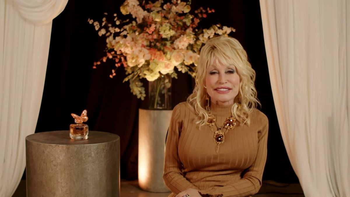 Dolly Parton sits smiling in a brown shirt beside a bottle of her perfume