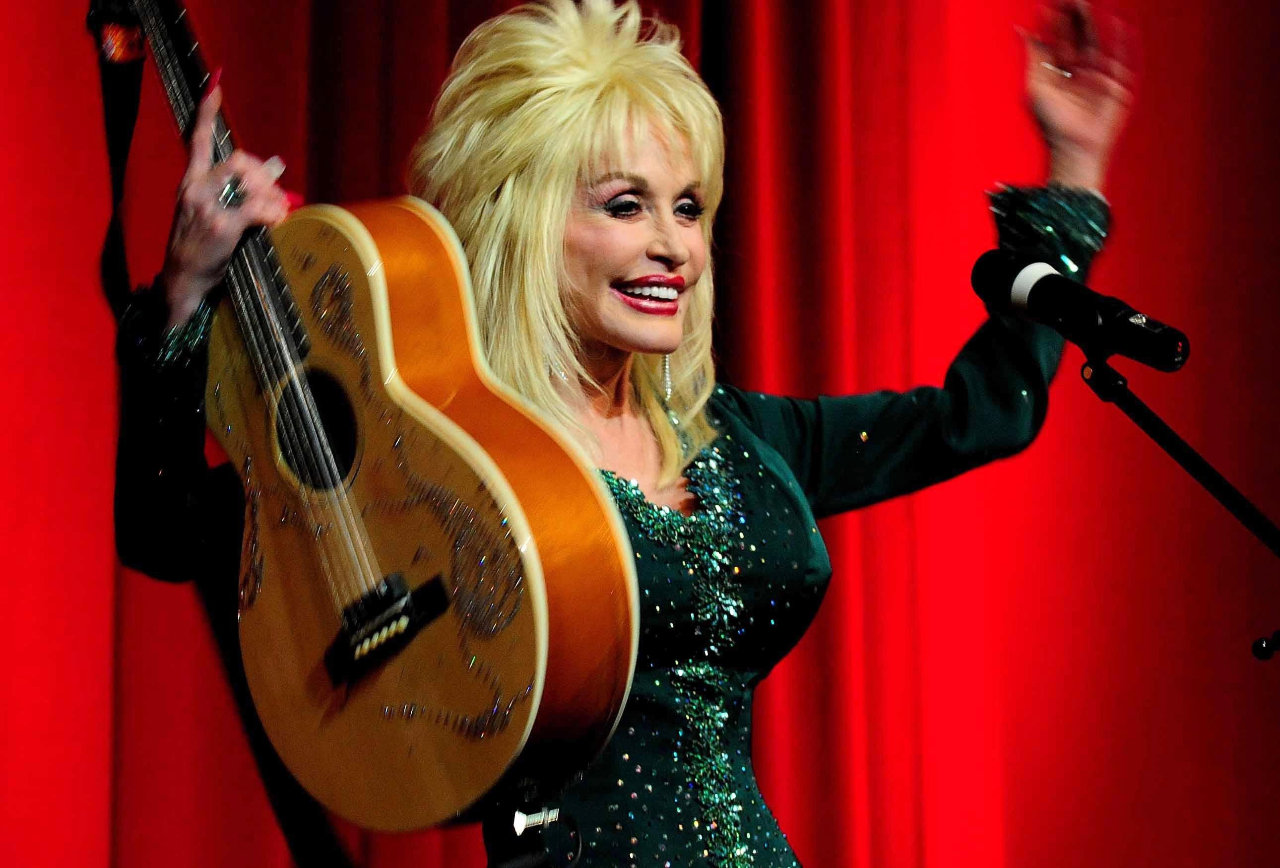 Dolly Parton holds up her hands and guitar in front of a red curtain.