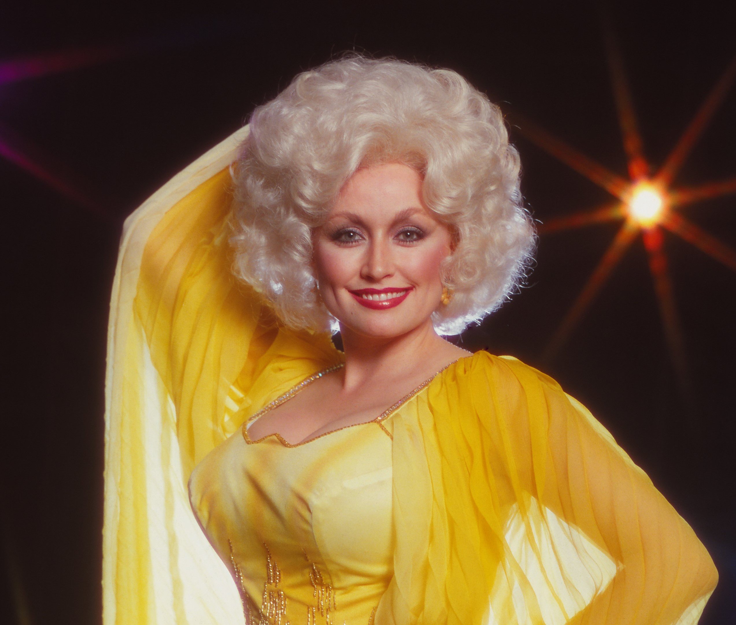 Dolly Parton wears a yellow dress in front of a black background.