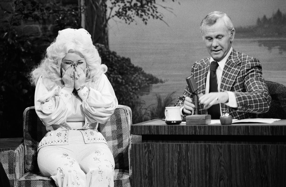 Dolly Parton covers her face and giggles while chatting with Johnny Carson, in a plaid jacket