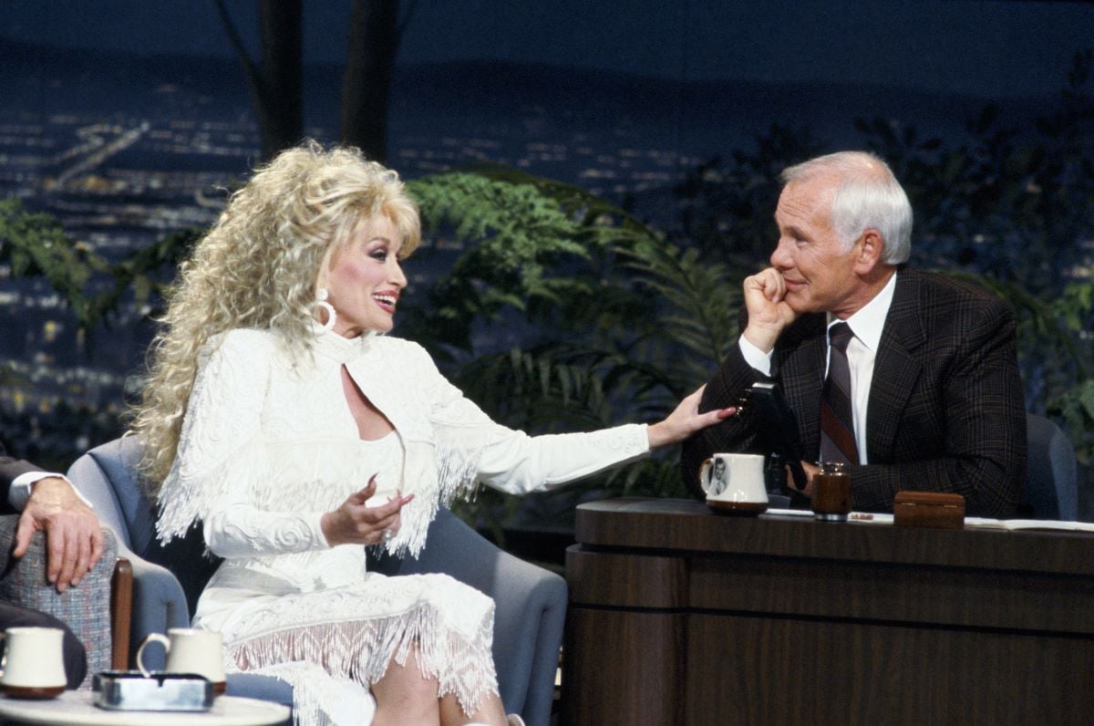 Guest Dolly Parton in white and host Johnny Carson in a suit and tie at his desk, leaning on his hand and looking at Parton