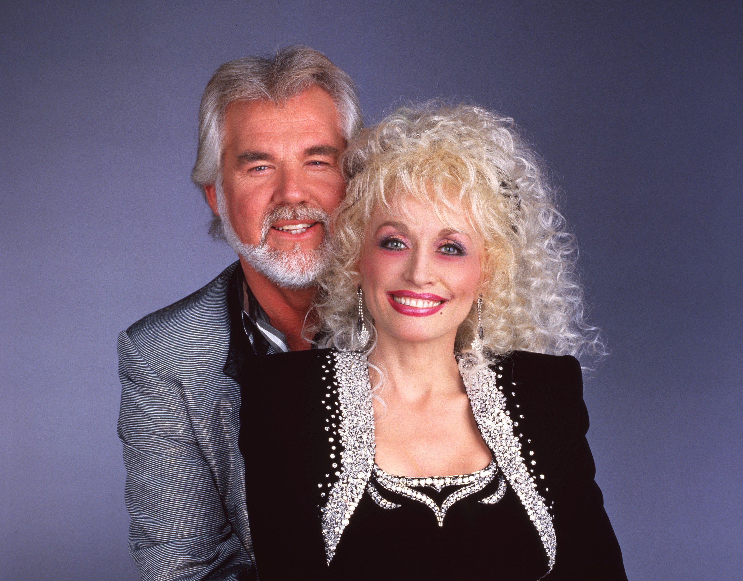 Kenny Rogers wears a gray shirt and stands behind Dolly Parton, who wears a black jacket and shirt.