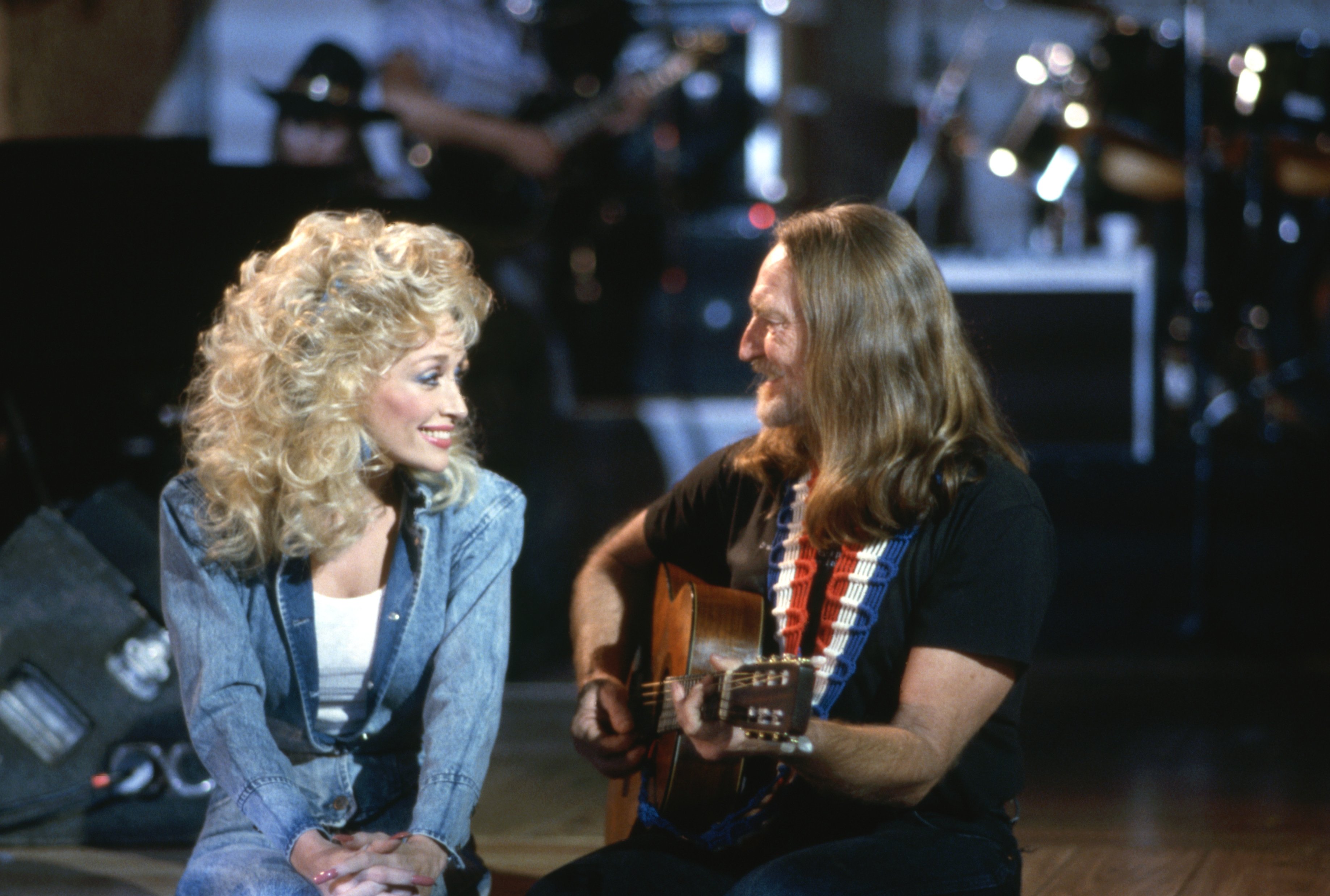 Dolly Parton wears a denim jacket and Willie Nelson wears a black shirt. He plays guitar.