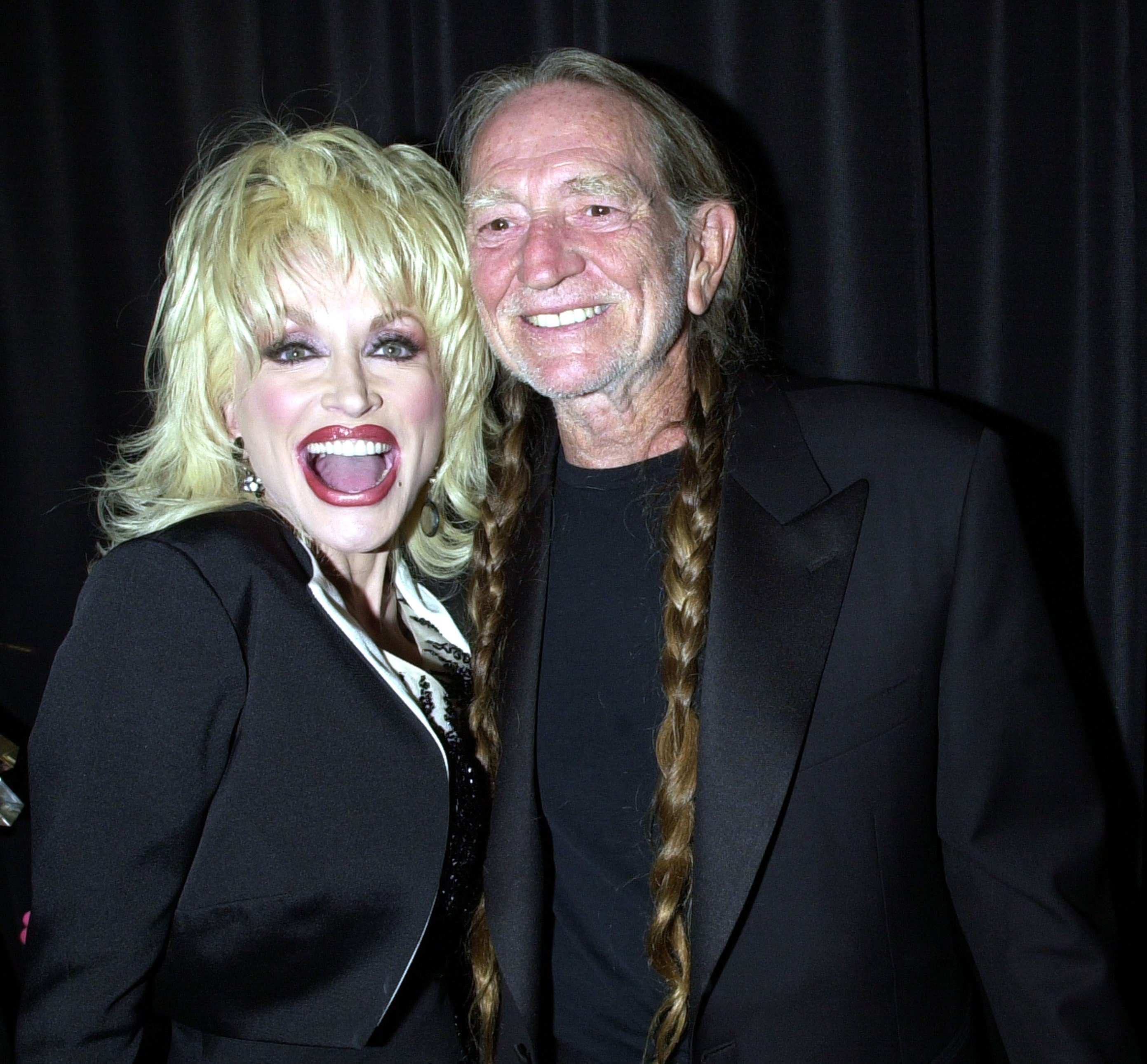 Dolly Parton and Willie Nelson both wear black outfits and stand in front of a black curtain.