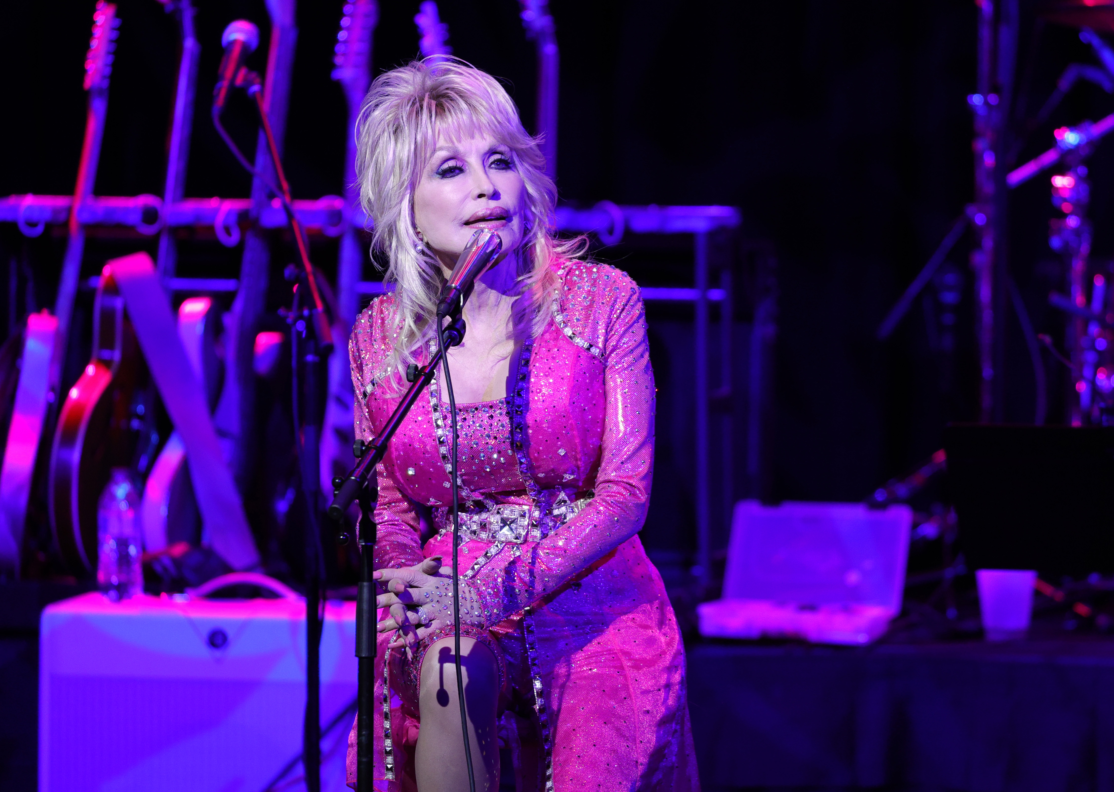 Dolly Parton sits in front of a microphone while dressed in a fuchsia outfit.