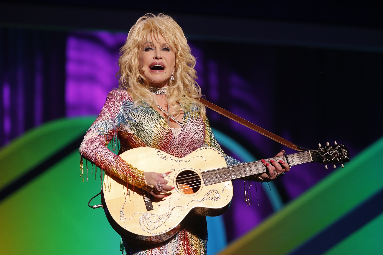 Dolly Parton wears a rainbow, sparkly shirt and strums a guitar.