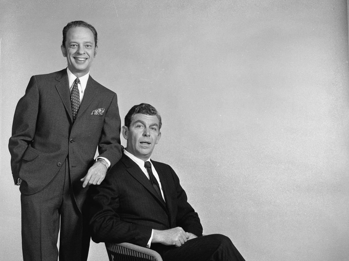 Don Knotts stands behind Andy Griffith, who is seated, and both are dressed in suits