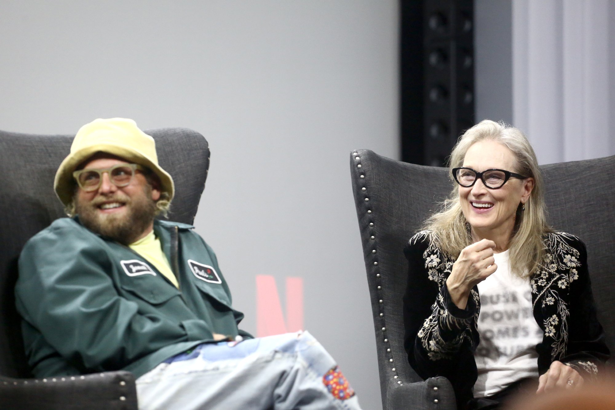 'Don't Look Up stars Meryl Streep and Jonah Hill smiling and sitting in grey couch chairs in front of a silver screen