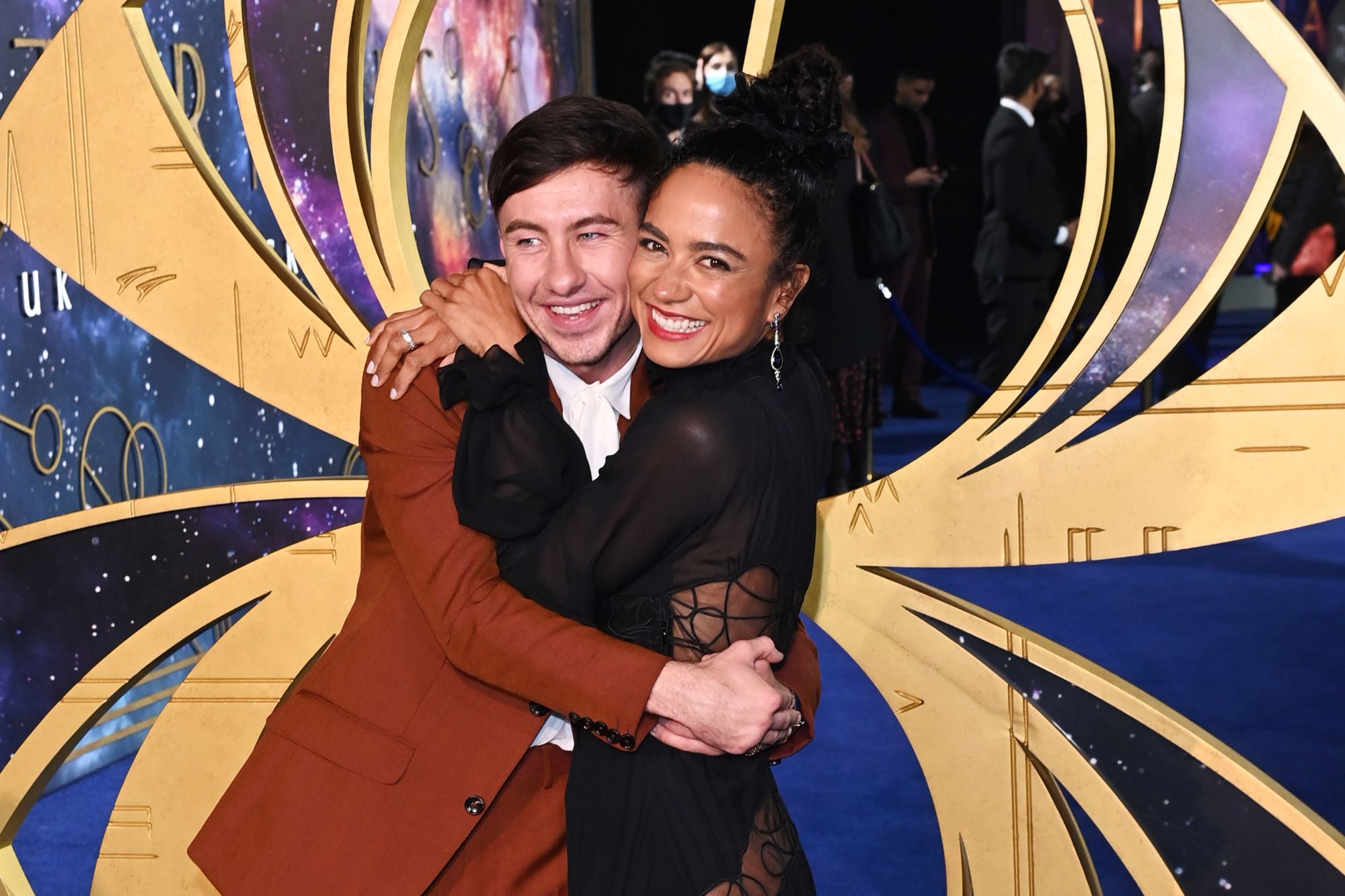 Marvel's 'Eternals' stars Barry Keoghan and Lauren Ridloff embrace one another on the red carpet. Keoghan wears a brown red suit and pants and a white shirt. Ridloff wears a black long-sleeved dress. Keoghan and Ridloff play Druig and Makkari in the film, a couple that many fans prefer over Sersi and Ikaris.