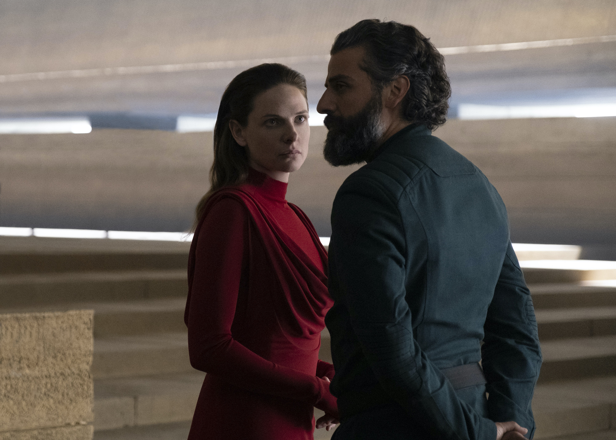 Rebecca Ferguson as Lady Jessica and Oscar Isaac as Duke Leto Atreides in 'Dune.' They stand in a room made of tan stone floors and walls with the sun peeking in behind them. Ferguson wears a red turtleneck dress with long sleeves. Isaac wears a dark faded blue jacket. They both look concerned.