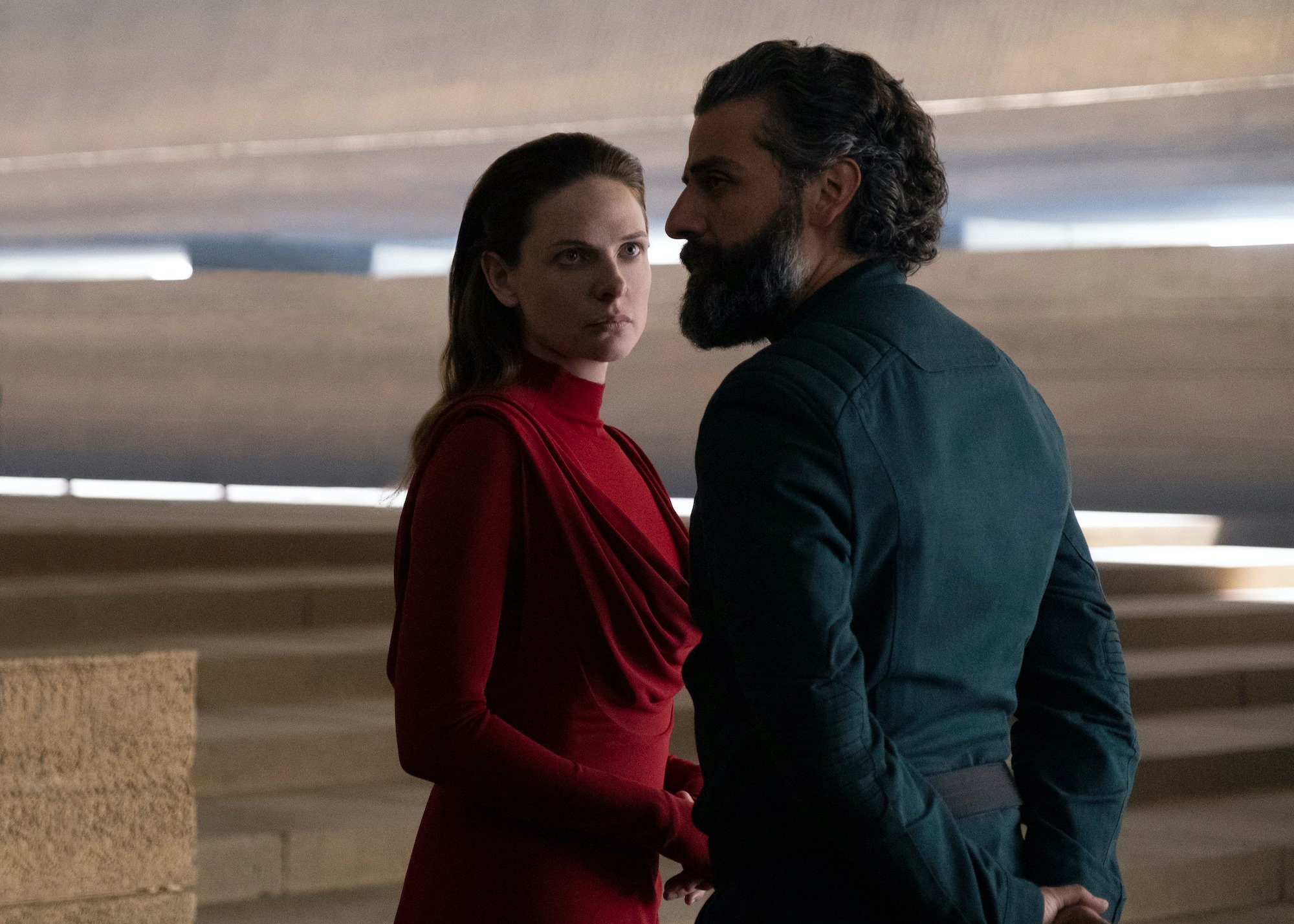 Rebecca Ferguson as Lady Jessica and Oscar Isaac as Duke Leto Atreides in 'Dune.' They stand in a room made of tan stone floors and walls with the sun peeking in behind them. Ferguson wears a red turtleneck dress with long sleeves. Isaac wears a dark faded blue jacket. They both look concerned.