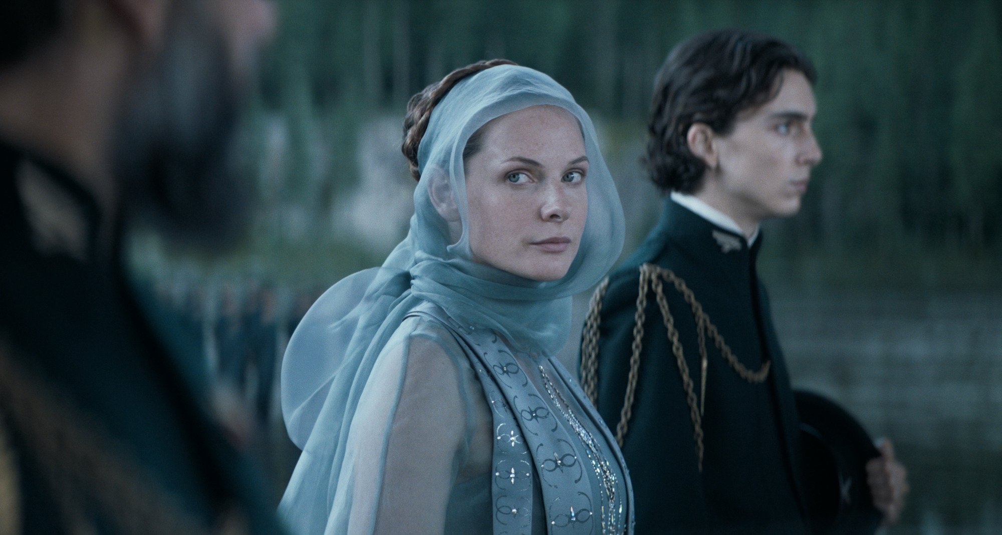 Rebecca Ferguson as Lady Jessica in 'Dune.' She wears a formal light blue gown with blue tulle around her head, neck, and shoulders. Timothée Chalamet stands in the background as Paul Atreides wearing a black formal jacket with a high neck and gold chains on his right shoulder.