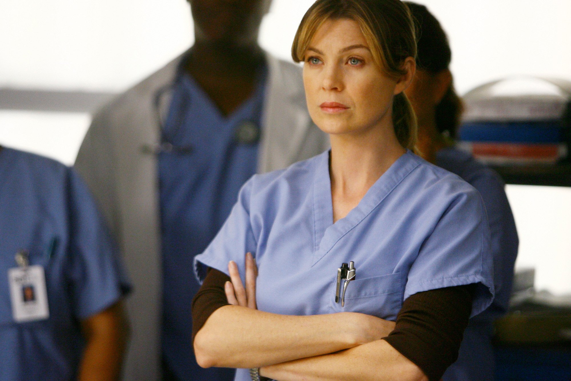 Ellen Pompeo as Meredith Grey in ABC's 'Grey's Anatomy.' She's wearing blue scrubs and her arms are crossed.