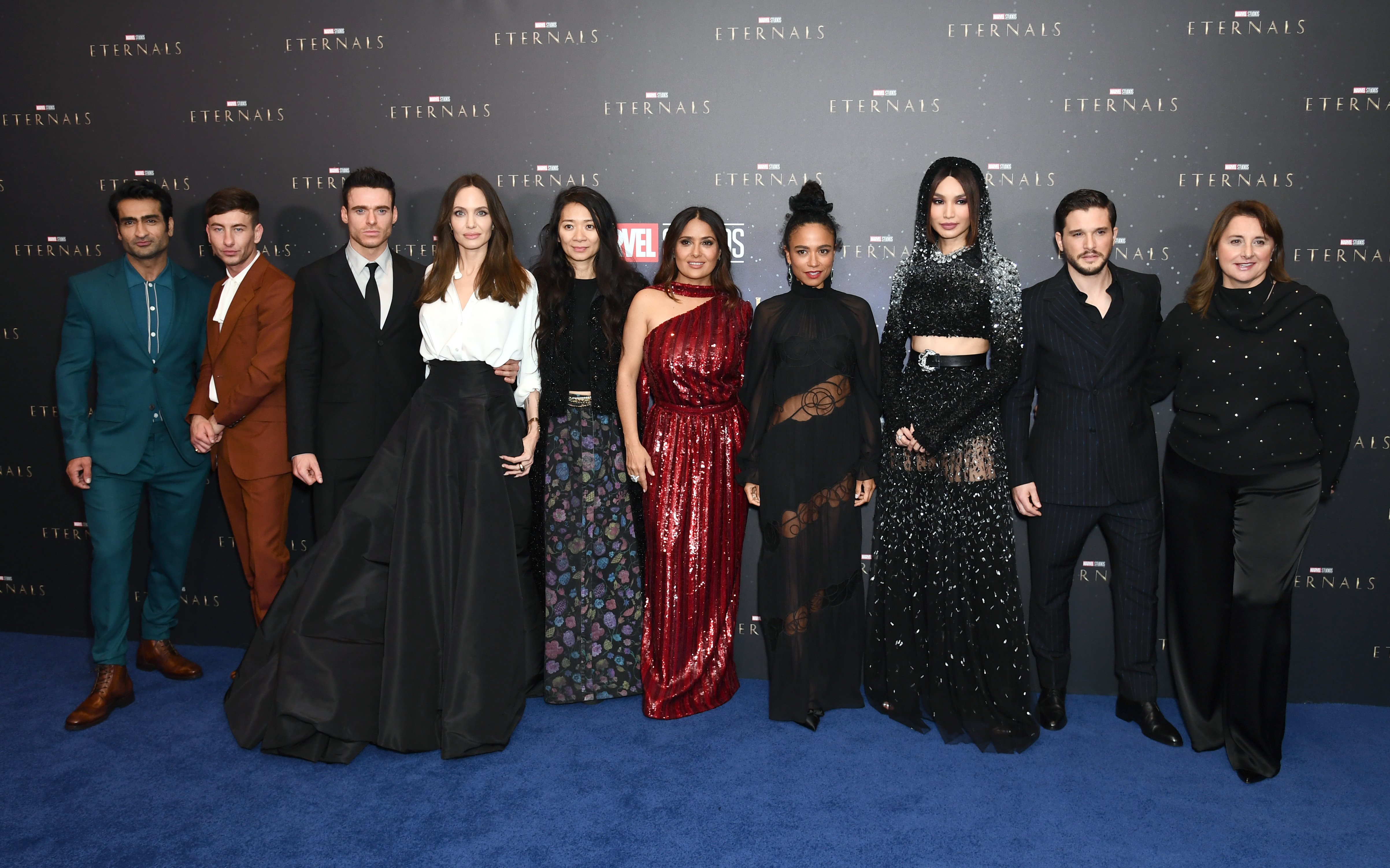 The cast and crew of Marvel's 'Eternals,' Kumail Nanjiani, Barry Keoghan, Richard Madden, Angelina Jolie, Chloe Zhao, Salma Hayek, Lauren Ridloff, Gemma Chan, Kit Harington, and Victoria Alonso, pose for a picture in front of a wall with the 'Eternals' logo on it. They are all dressed formally, in suits and dresses.