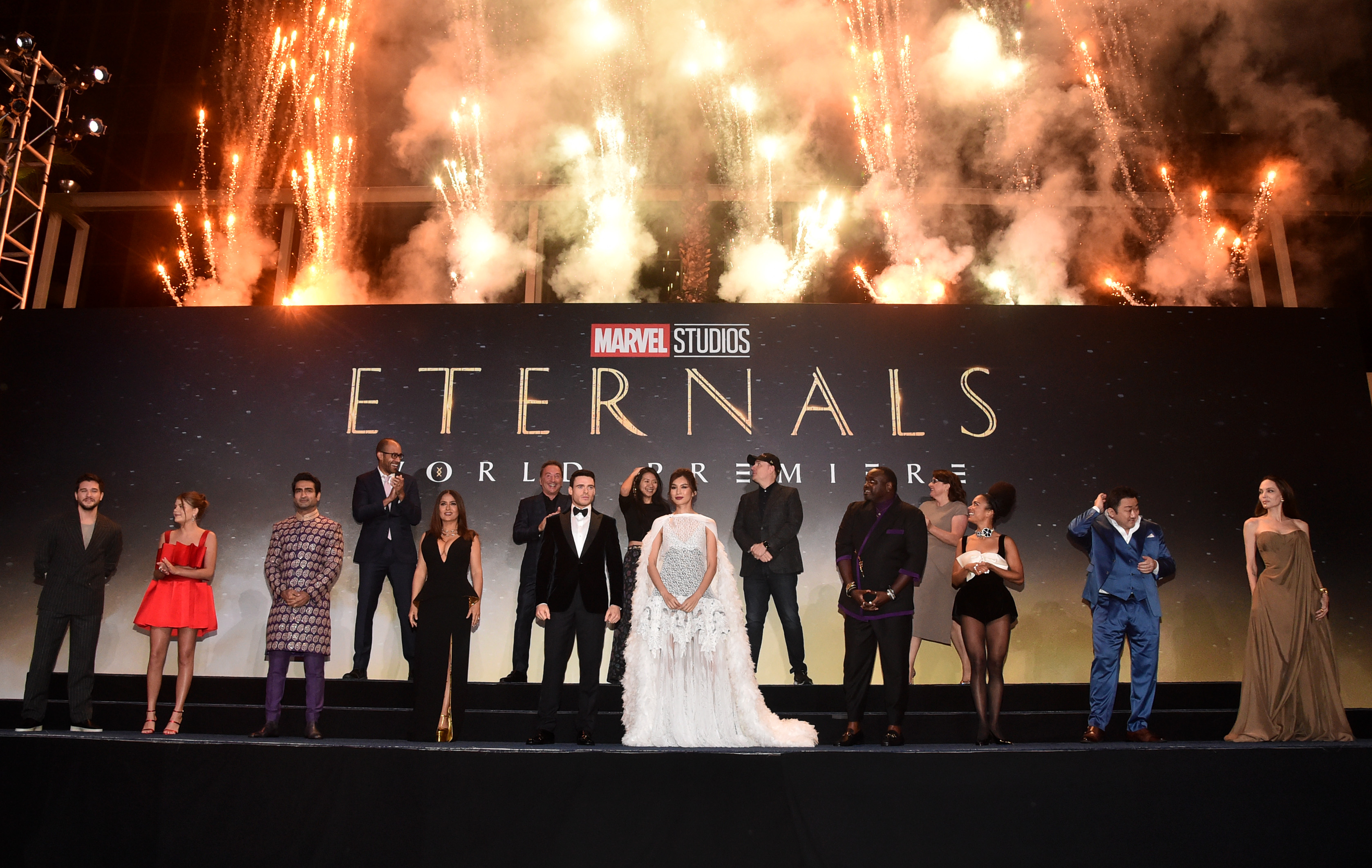 The cast and crew of Marvel's 'Eternals,' including director Chloé Zhao, pose for pictures in front of a poster for the film as fireworks go off behind them. 