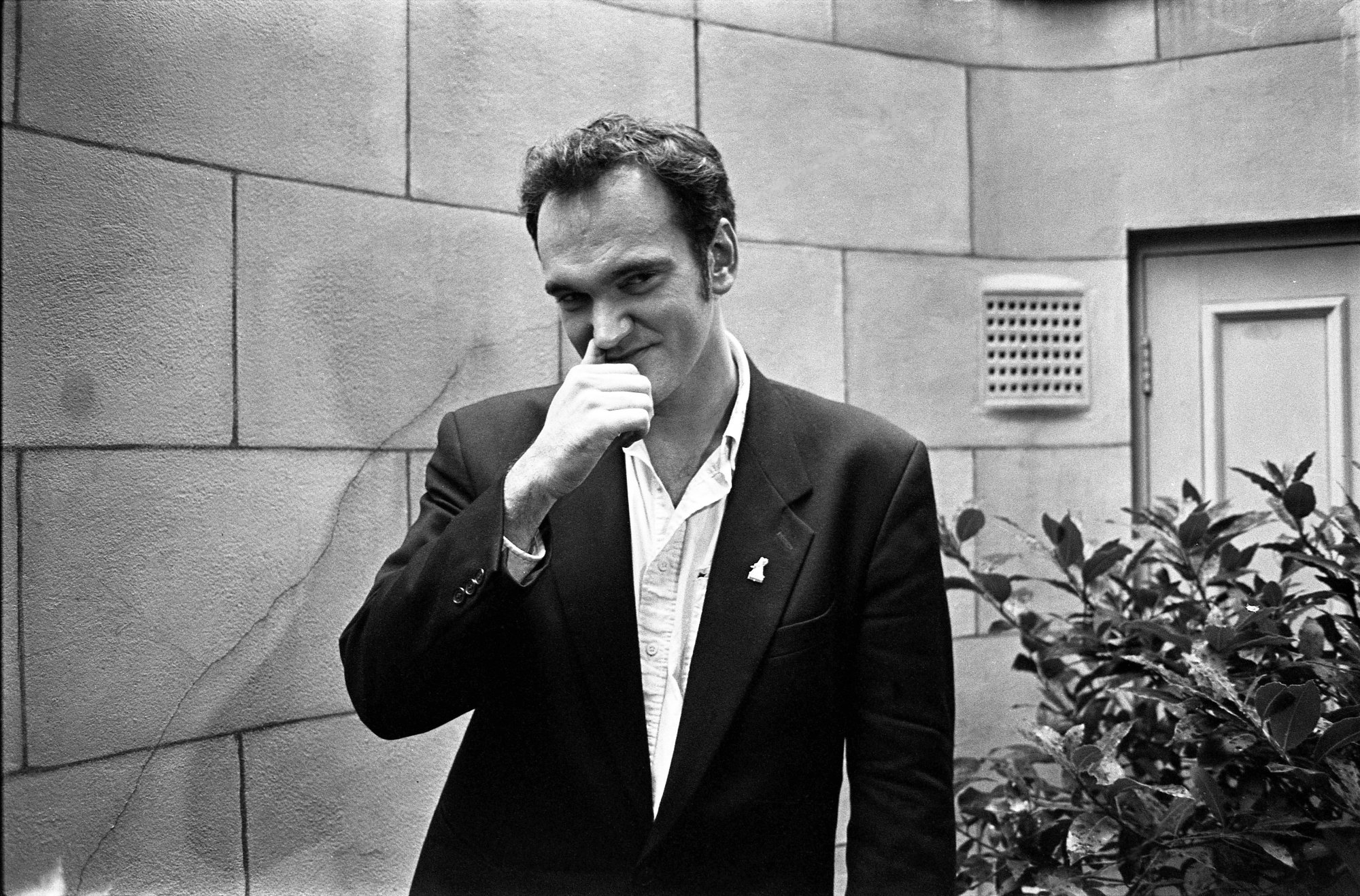 Quentin Tarantino in black-and-white wearing, wearing a suit with his finger on his nose. A featured image for an article about Quentin Tarantino's first-ever screenplay