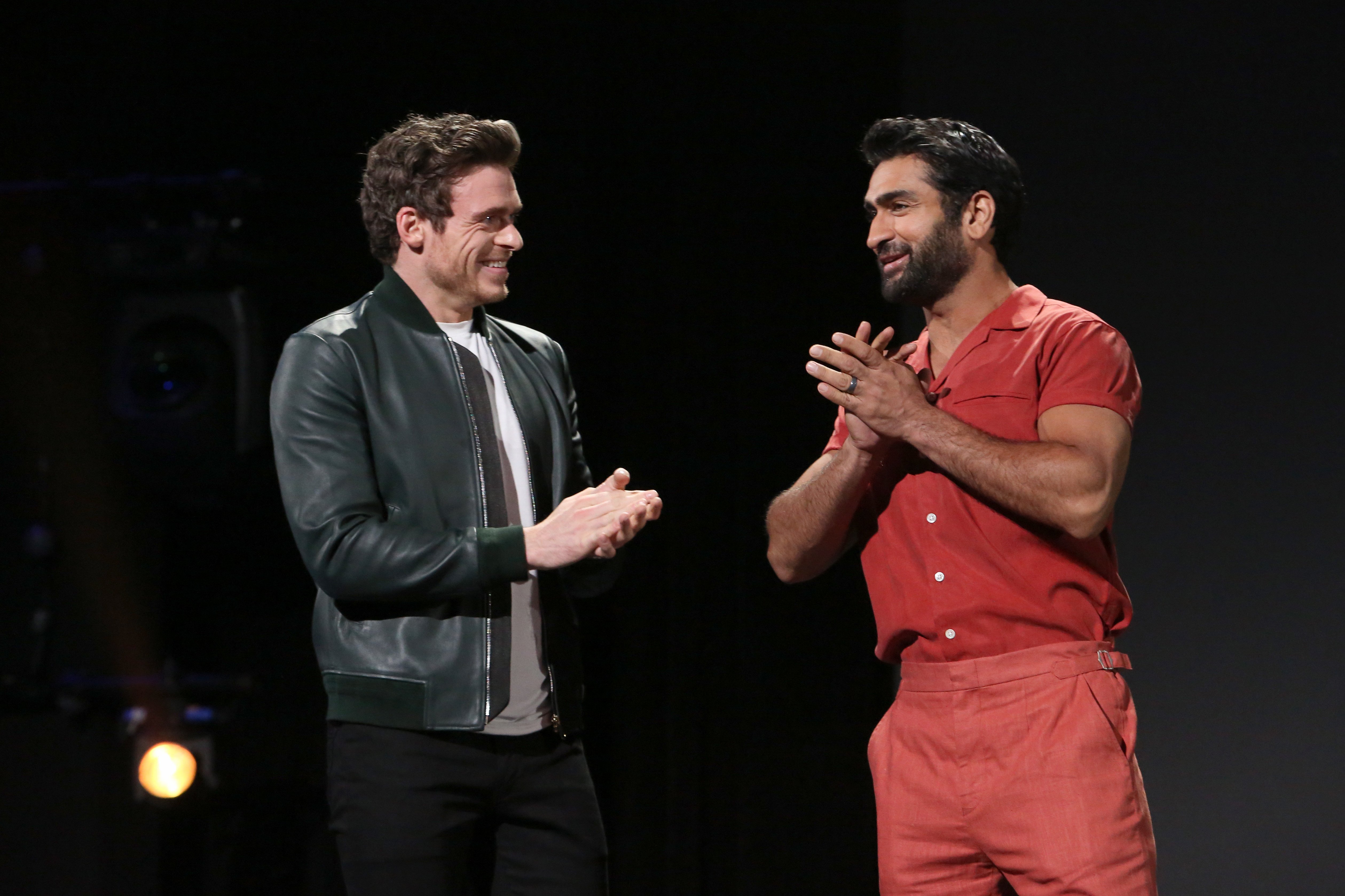 Marvel's 'Eternals' stars Richard Madden and Kumail Nanjiani stand side-by-side onstage. Madden wears a dark green leather jacket over a white shirt and black pants. Nanjiani wears a short-sleeved button-up red shirt and pink pants.