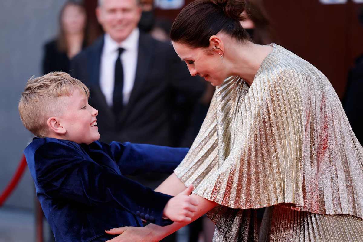 Jude Hill (L) and Caitriona Balfe (R) embrace on the red carpet of the European premiere of 'Belfast', during the 2021 BFI London Film Festival in London on Oct. 12, 2021. Hill wears a blue velvet blazer and Balfe wears a gold shimmering dress. They reach out for a hug.