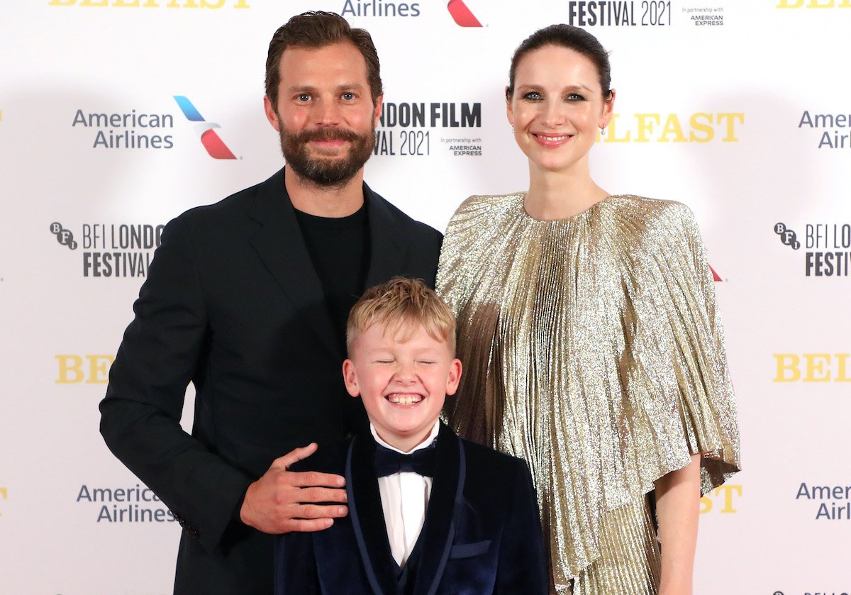 The cast of 'Belfast': Jamie Dornan, Jude Hill, and Caitriona Balfe attend the 'Belfast' European Premiere during the 65th BFI London Film Festival at The Royal Festival Hall on Oct. 12, 2021 in London, England. 11-year-old Hill wears a blue velvet blazer and smiles wide with his eyes closed. Dornan's hand is on Hill's right shoulder as he stands behind him smirking, and he wears a black suit and black crew-neck shirt. Balfe stands next to Dornan smiling while wearing a gold shimmering dress.