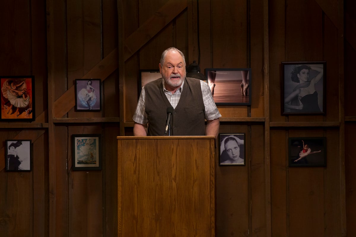 Michael Winters as Taylor Doose behind a podium at a Stars Hollow town meeting