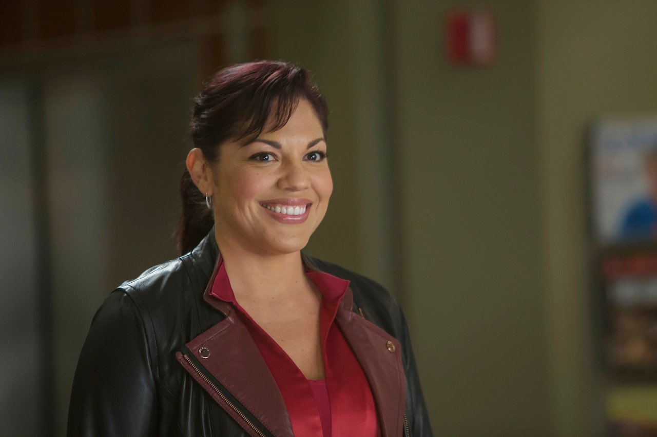 Sara Ramirez as Callie Torres on 'Grey's Anatomy' smiles wearing a red top and brown jacket.