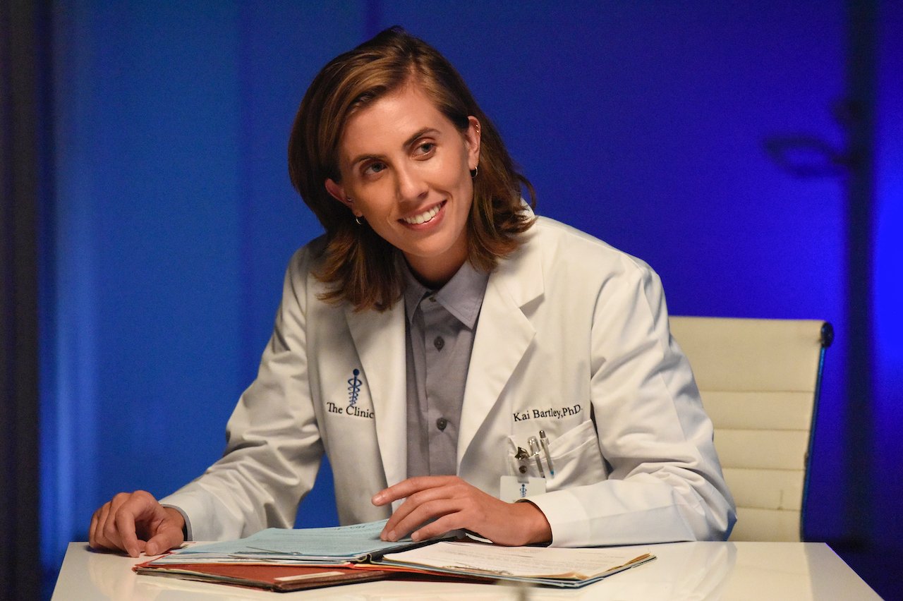 E. R. Fightmaster as Dr. Kai Bartley sits at a desk in a lab coat smiling.