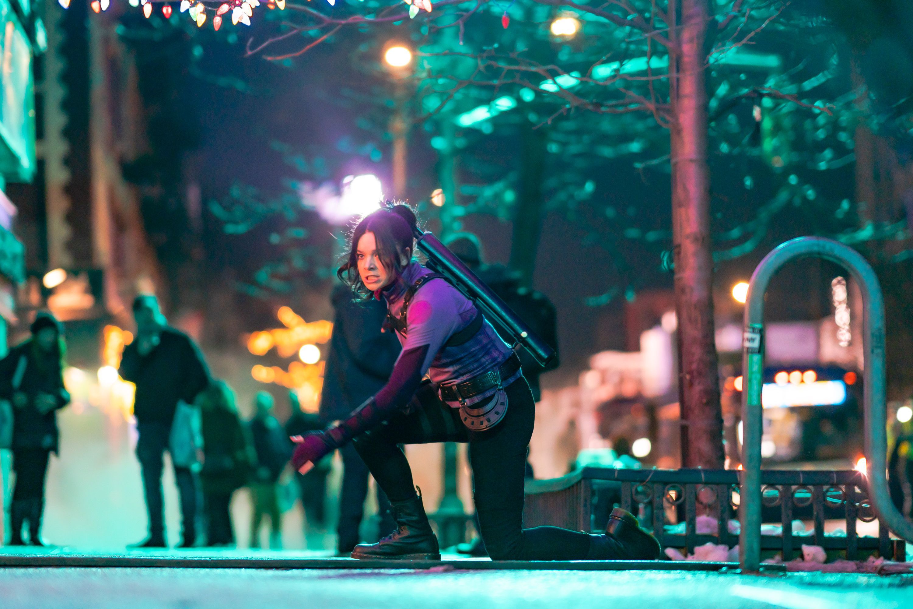 'Hawkeye' actor Hailee Steinfeld films scenes as Kate Bishop in New York City. Steinfeld wears a purple long-sleeved shirt, black pants, and black boots. She has a quiver slung around her shoulder as she kneels on the ground.