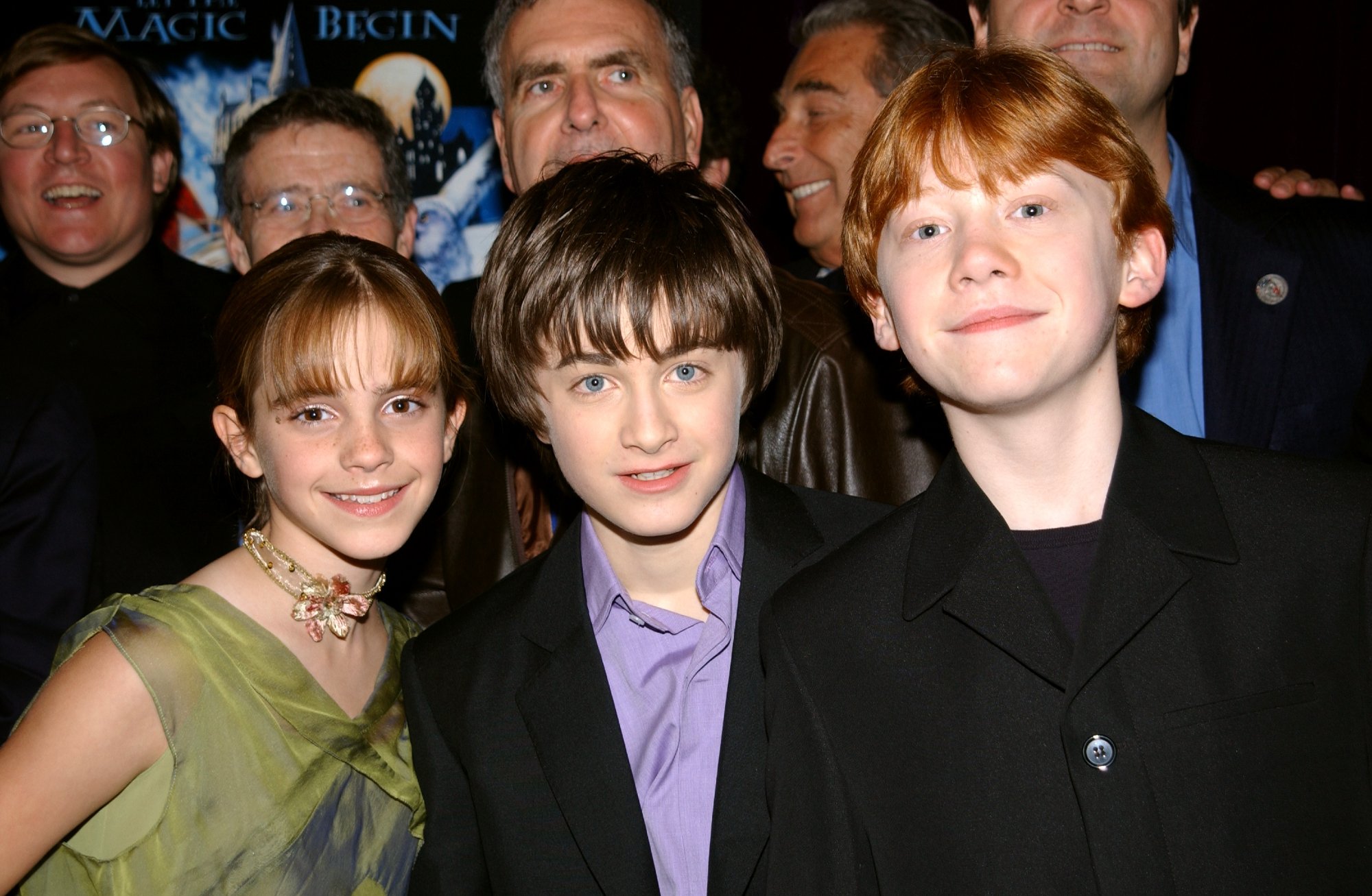 'Harry Potter' movies actors Emma Watson, Daniel Radcliffe, and Rupert Grint smiling in front of a 'Harry Potter' poster and a crowd of people